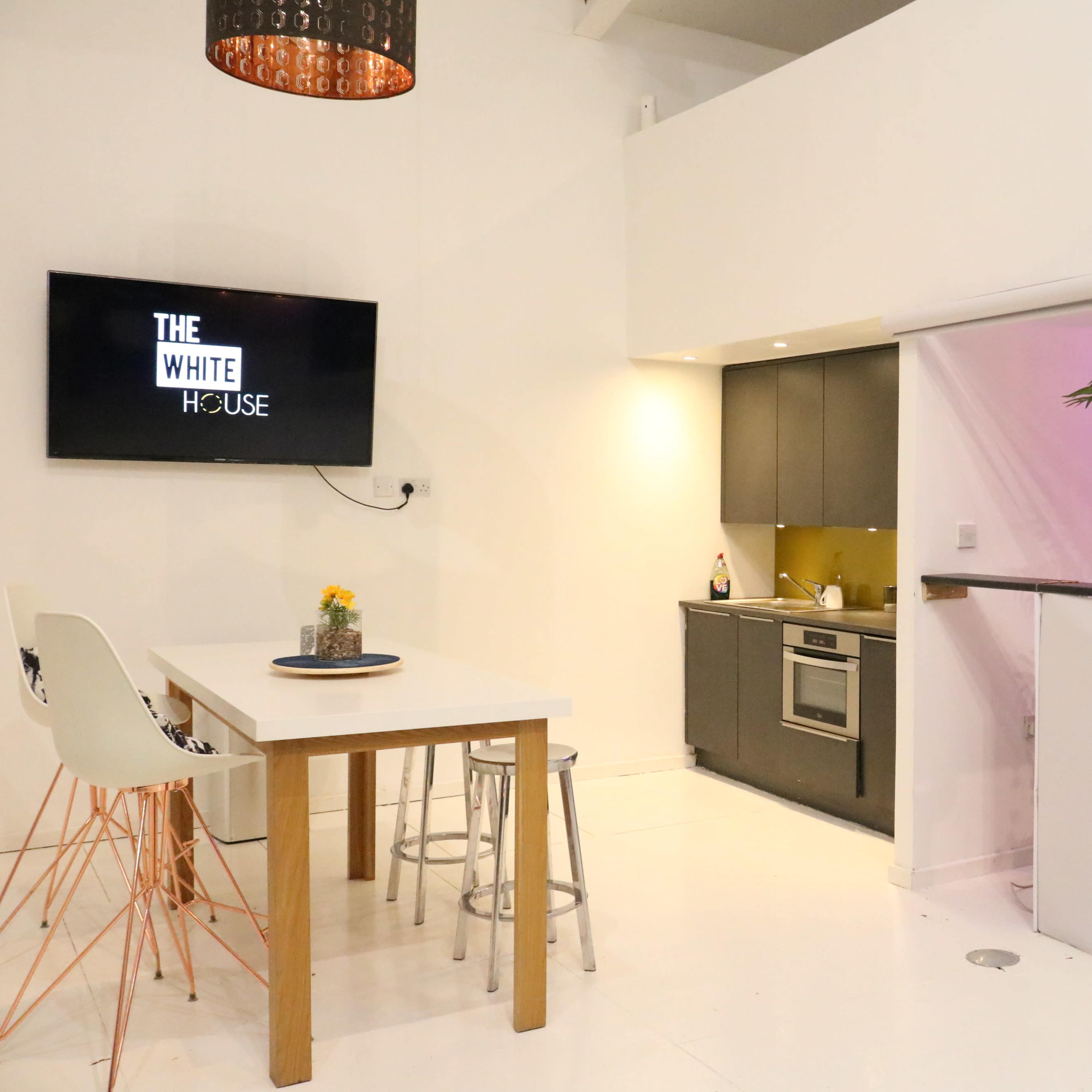 A modern kitchen and dining area featuring a white table with bar stools, a television on the wall displaying "THE WHITE HOUSE," and a counter with vibrant lighting and greenery.