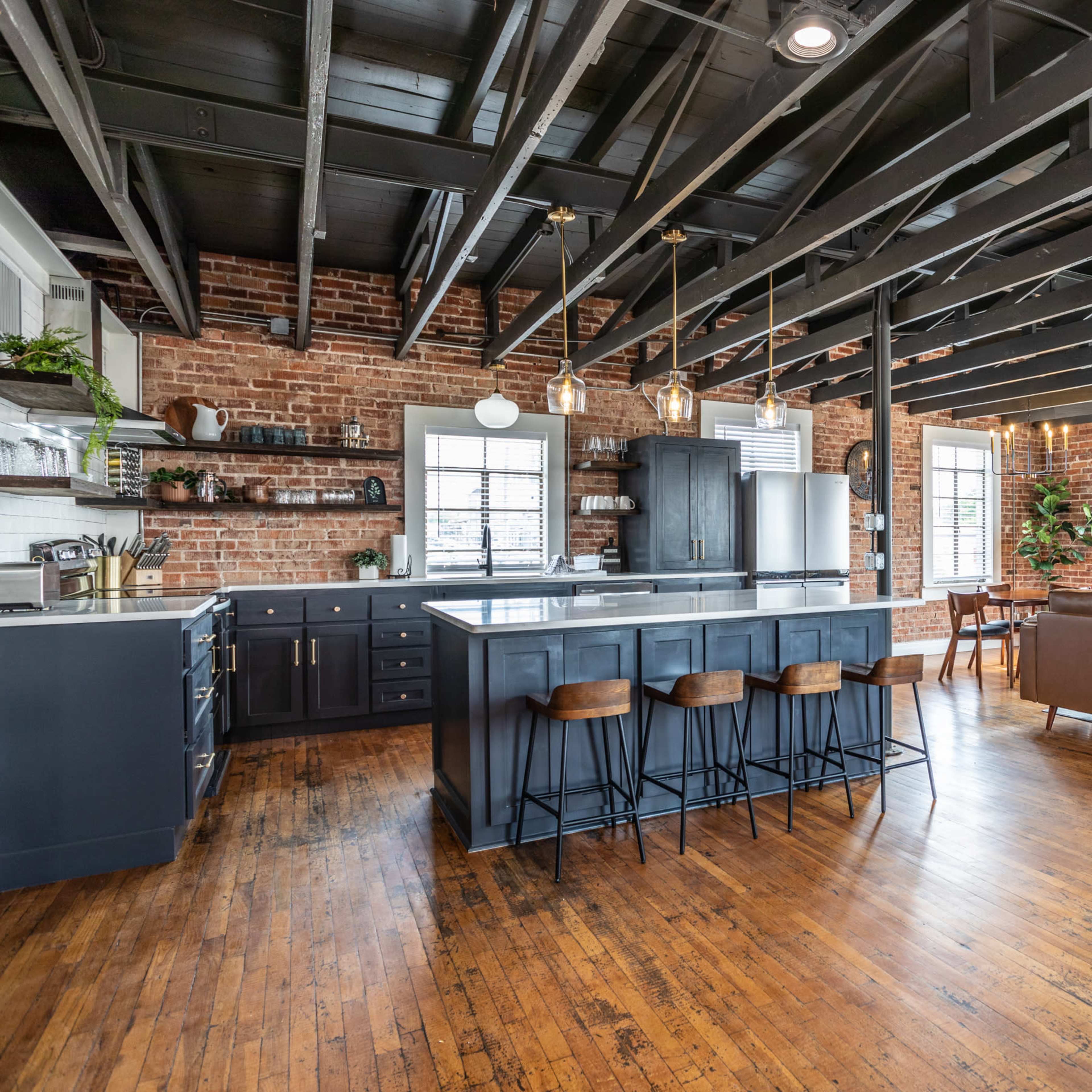 A modern kitchen and living area featuring exposed brick walls, a large island with bar stools, and a combination of dark cabinetry and wooden flooring.