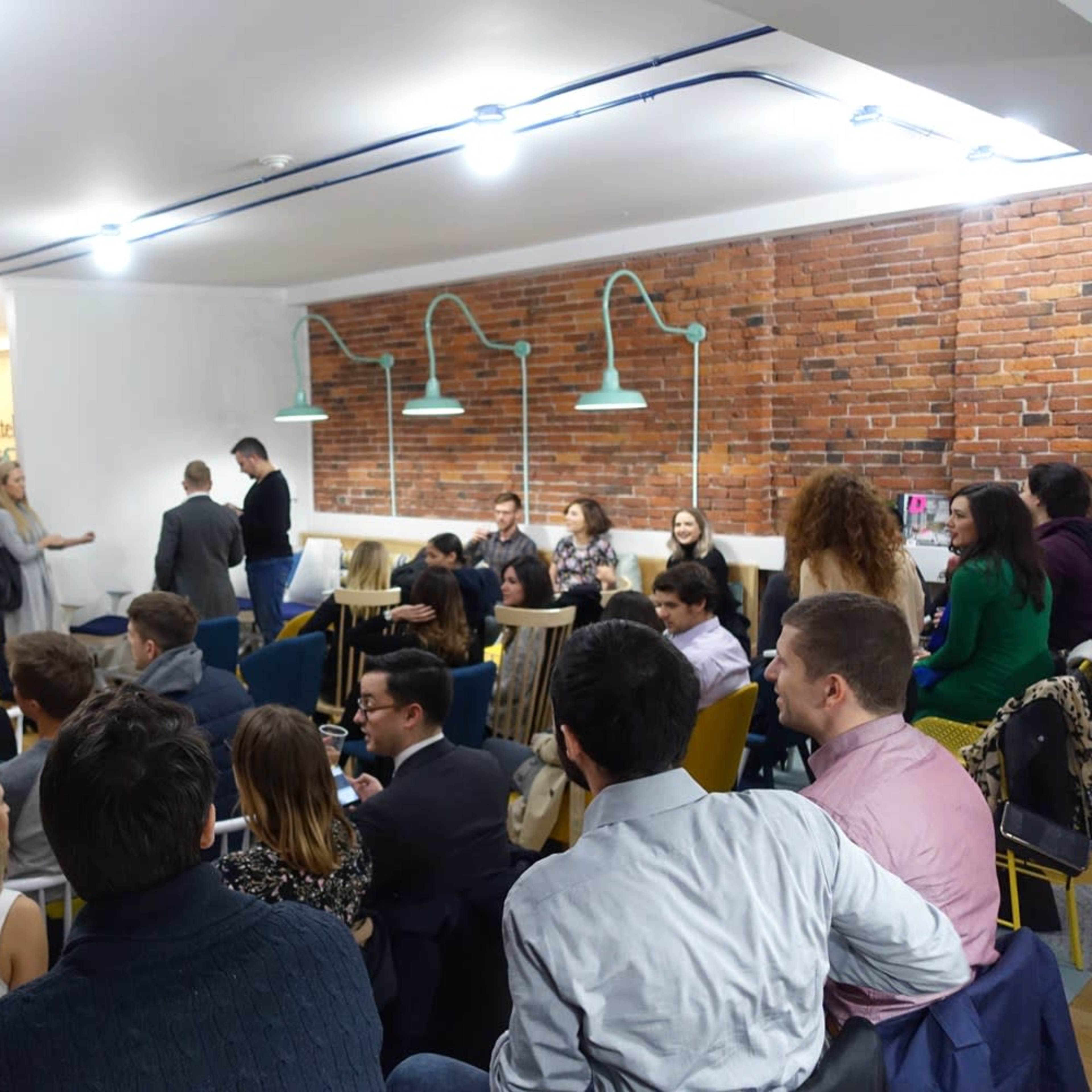 A group of people is seated and standing in a modern conference space with exposed brick walls and overhead lighting, engaged in conversation and watching a presentation.