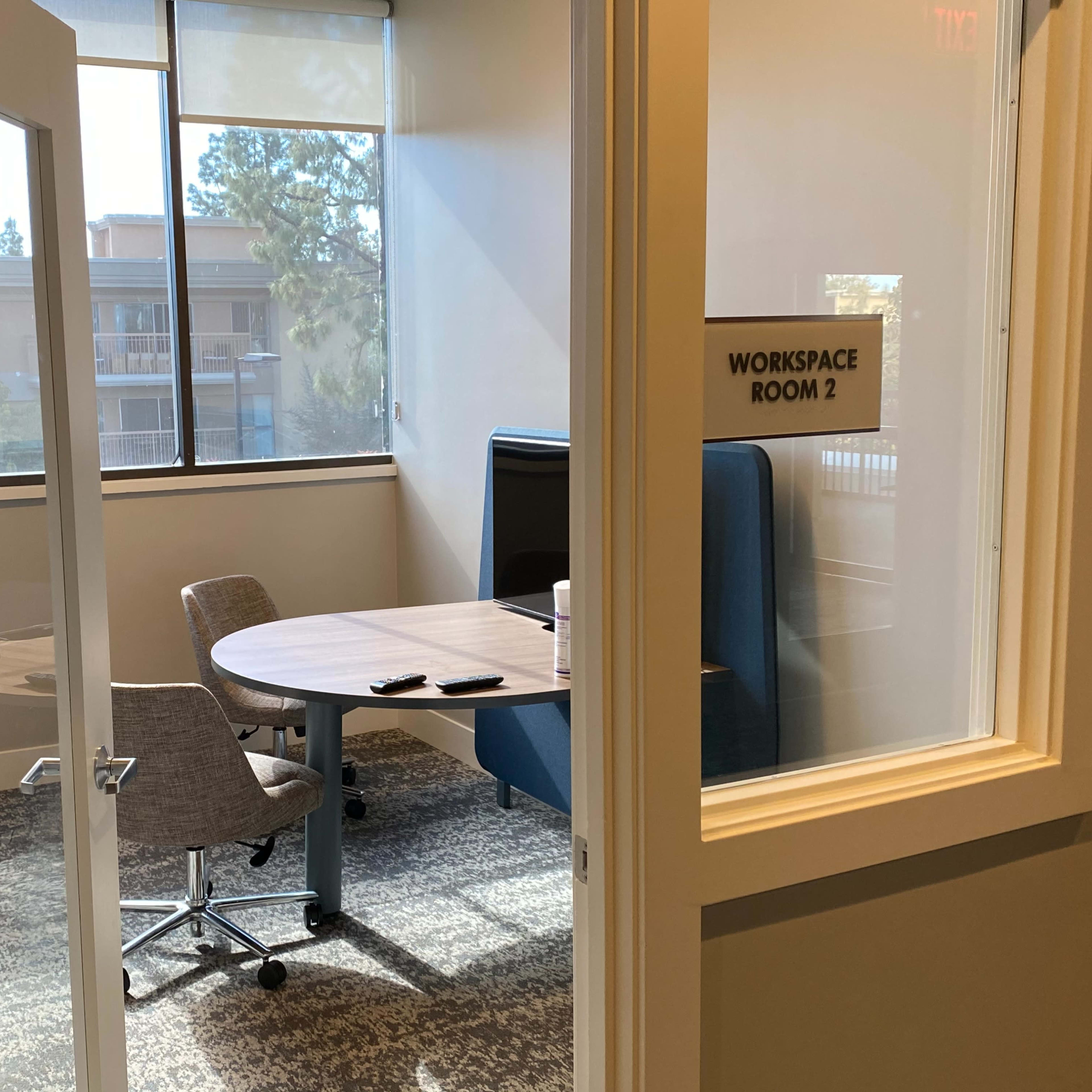 A small workspace with a round table and two chairs, visible through a glass door, in a well-lit room.