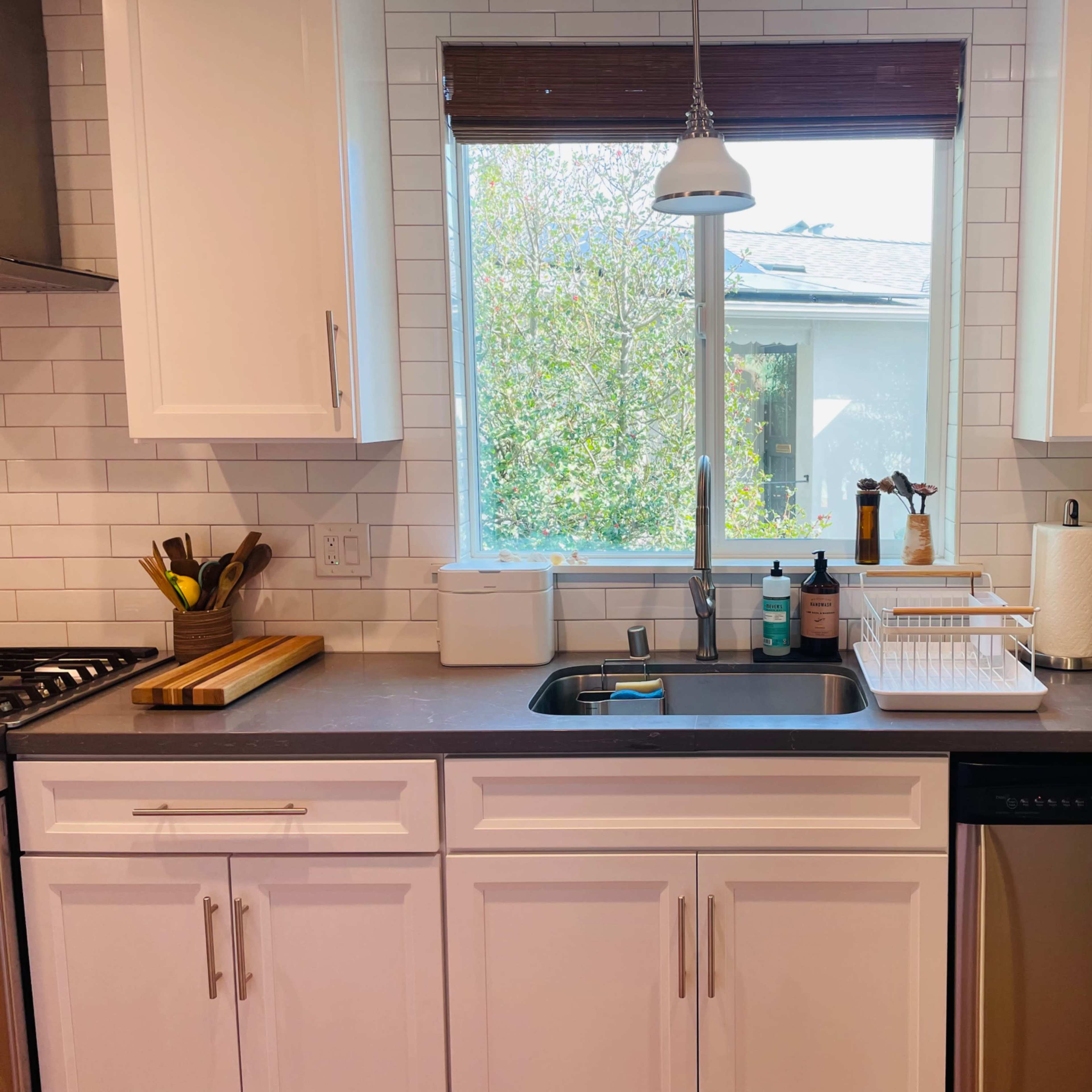 A kitchen countertop features a sink, a dishwasher, cutting boards, and various kitchen utensils, with a window providing natural light and a view of greenery outside.