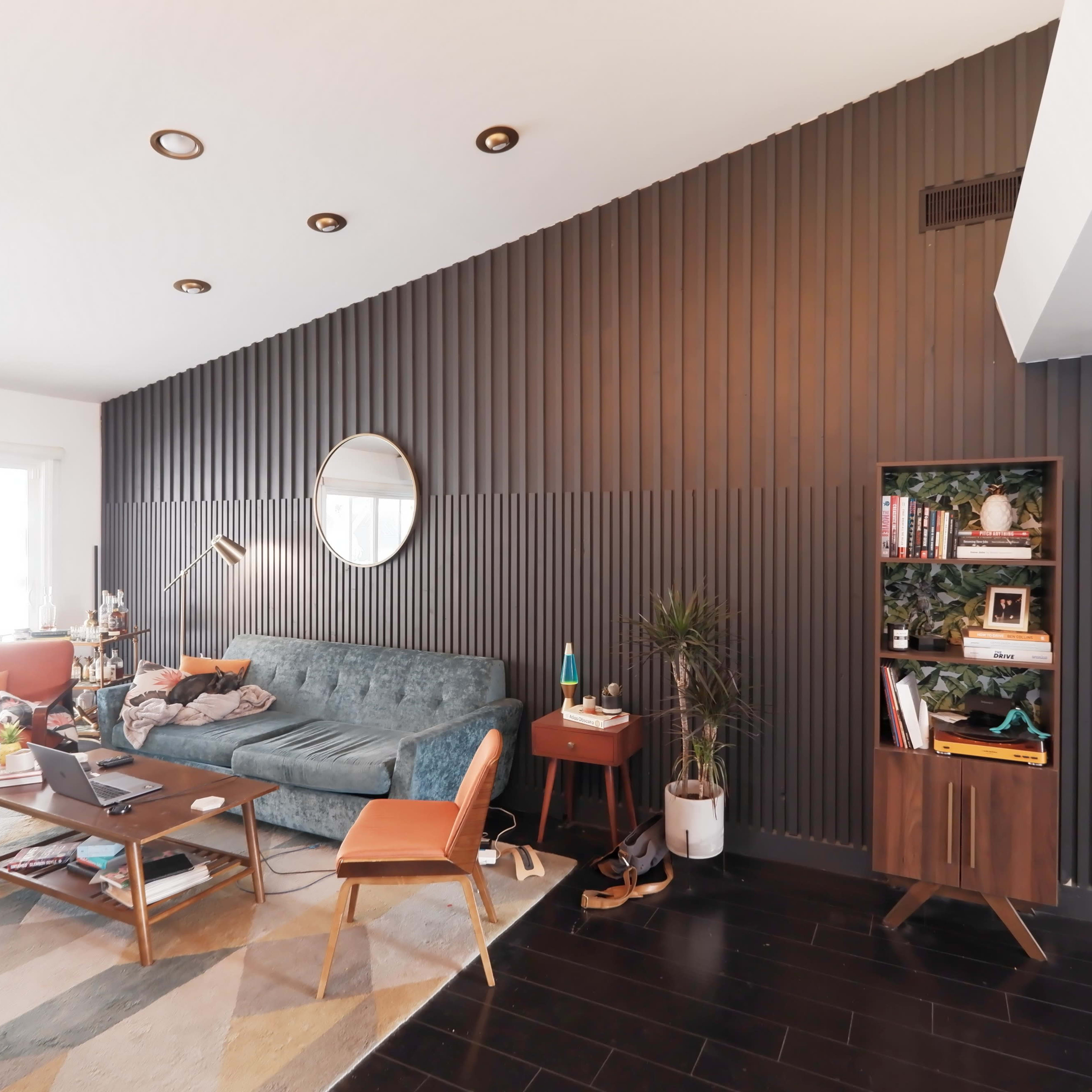A modern living room featuring a teal sofa, a wooden coffee table, a bookshelf, and a dark paneled wall with a round mirror.