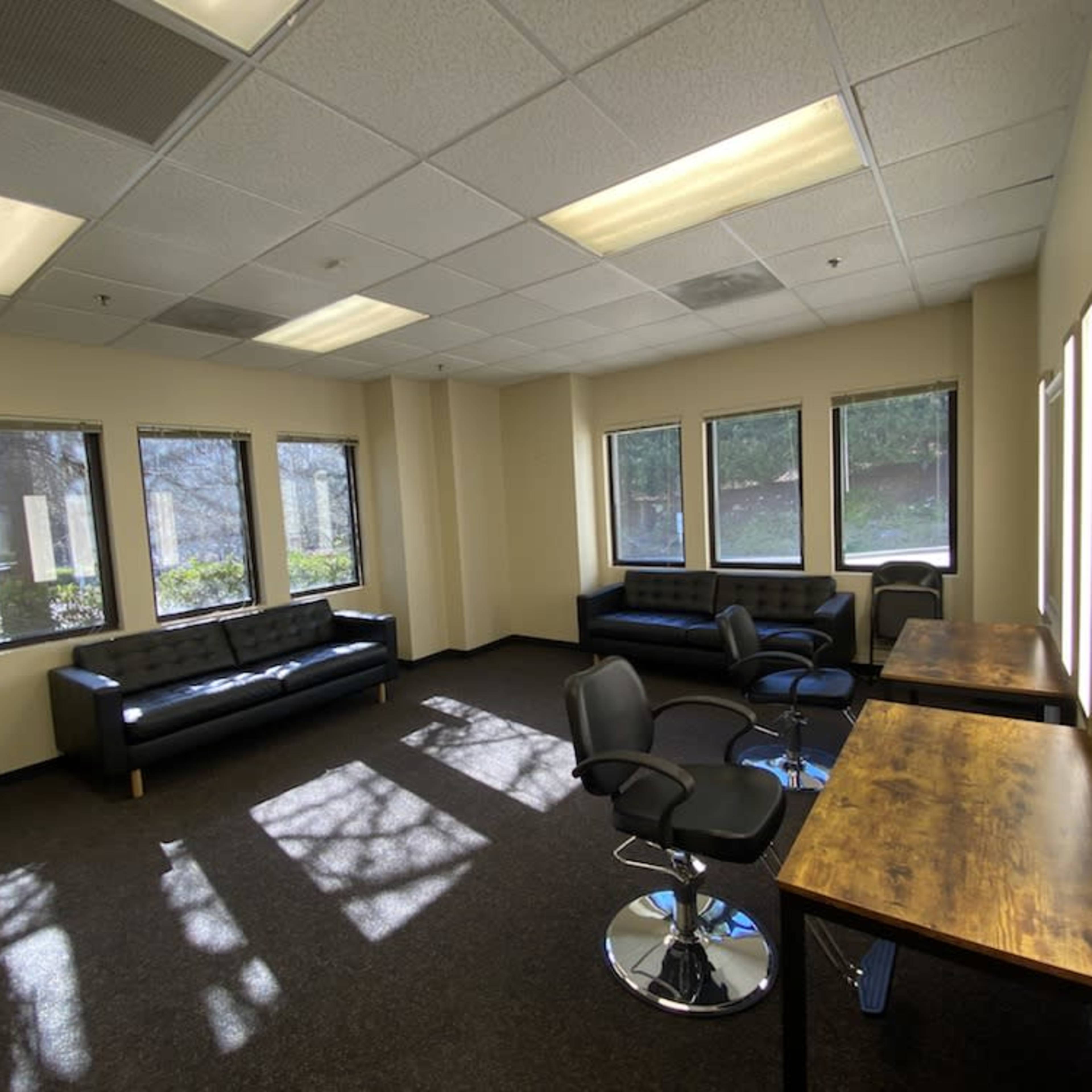 A room with two black sofas, several swivel chairs, and wooden tables, illuminated by natural light from large windows.