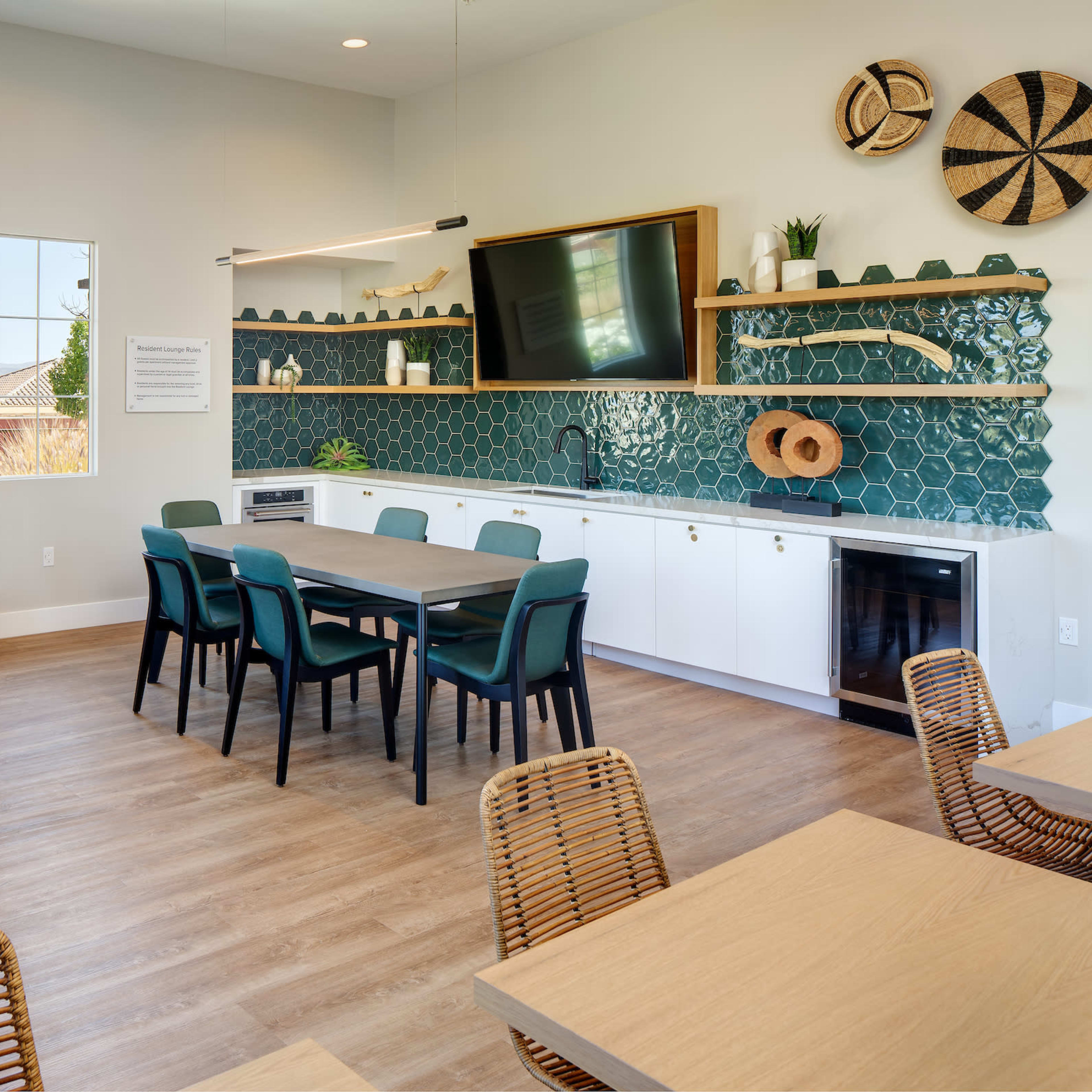 A bright, modern lounge area features a sofa, a dining table with chairs, and a kitchen area with green tile and decorative wall items.