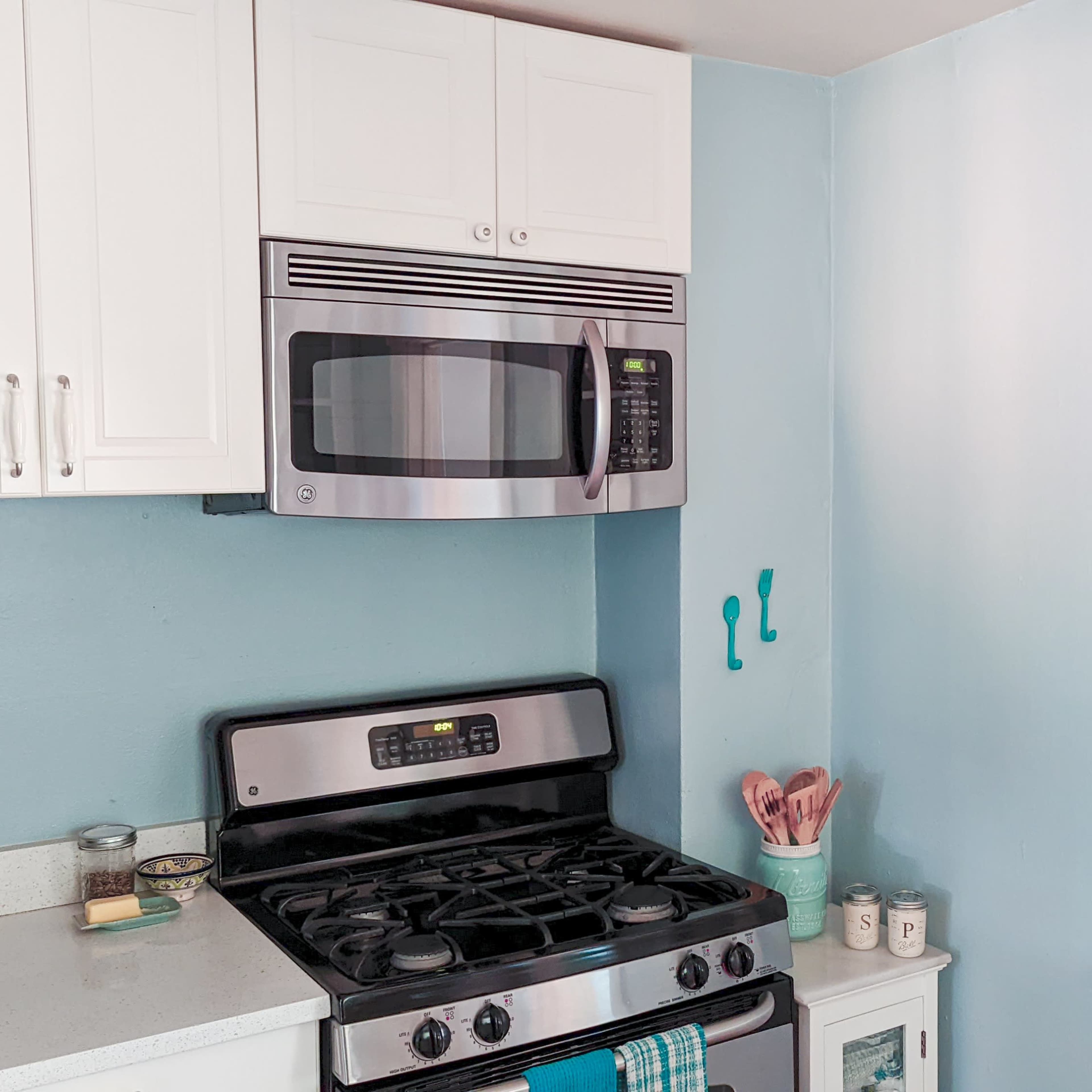 A modern kitchen features a stainless steel stove and microwave against light blue walls, with white cabinetry and shelves displaying various kitchen items.