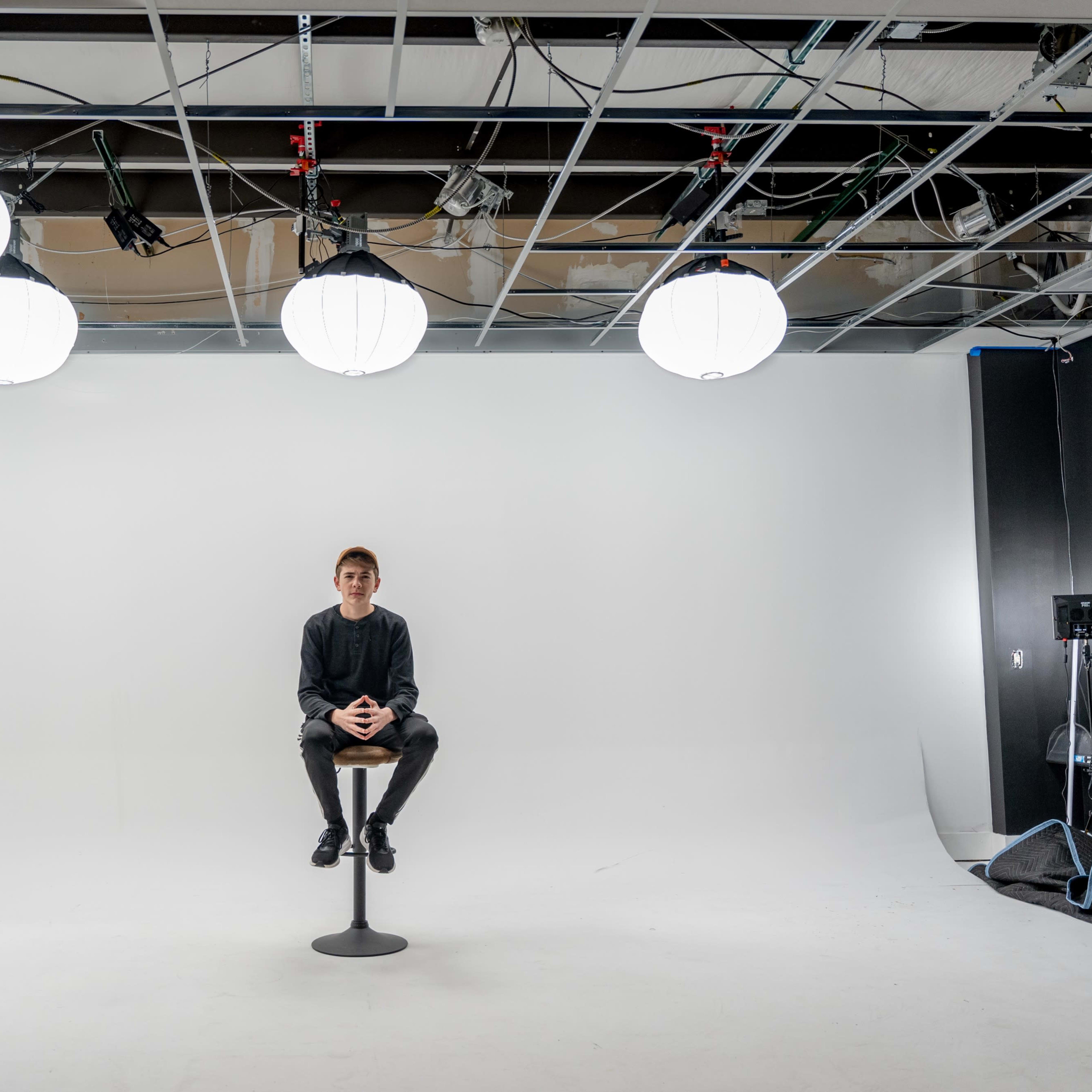 A person sits on a stool in a minimalist photo studio with bright pendant lights overhead and a plain white backdrop.