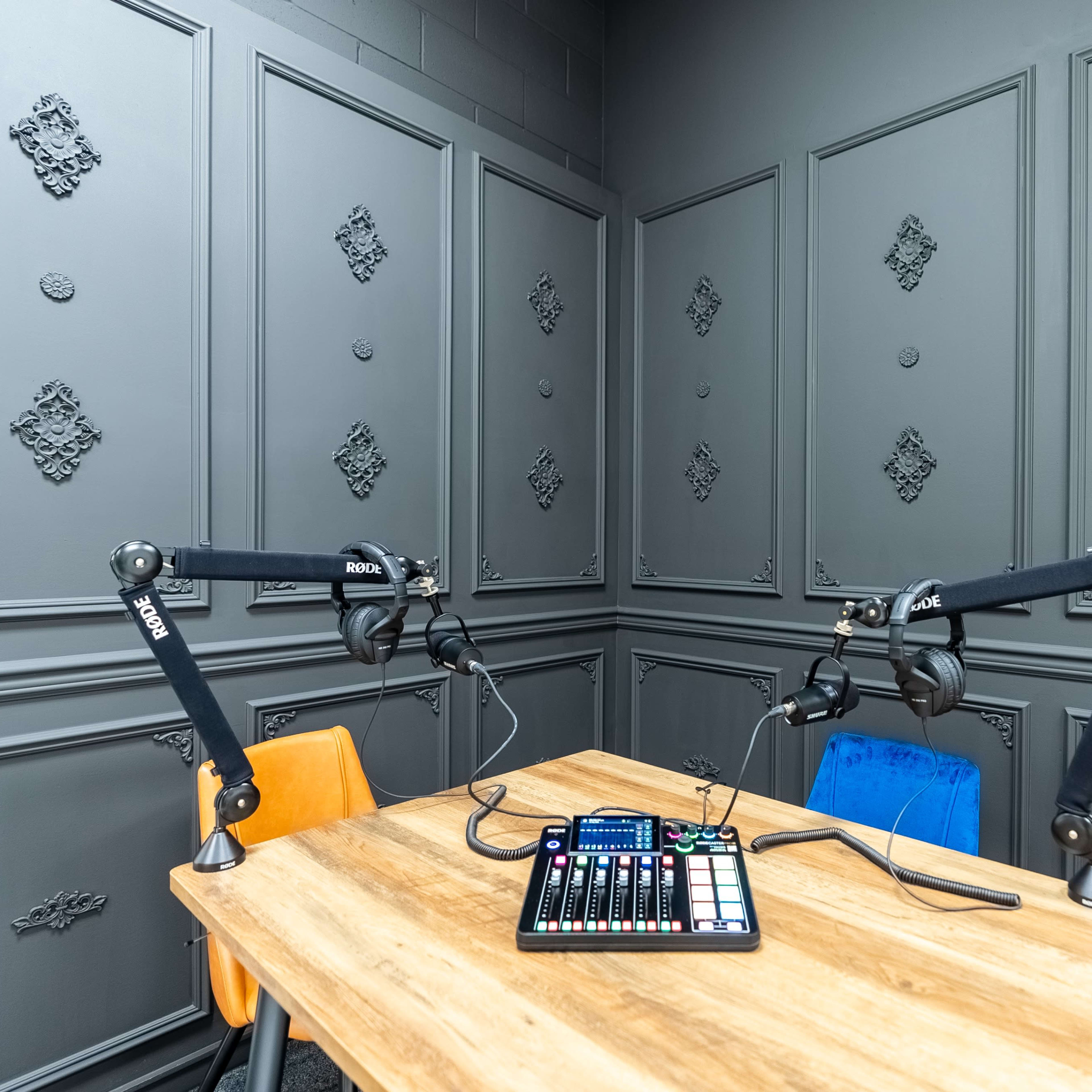 A recording studio features a wooden table with audio equipment and two microphone stands against textured gray walls.