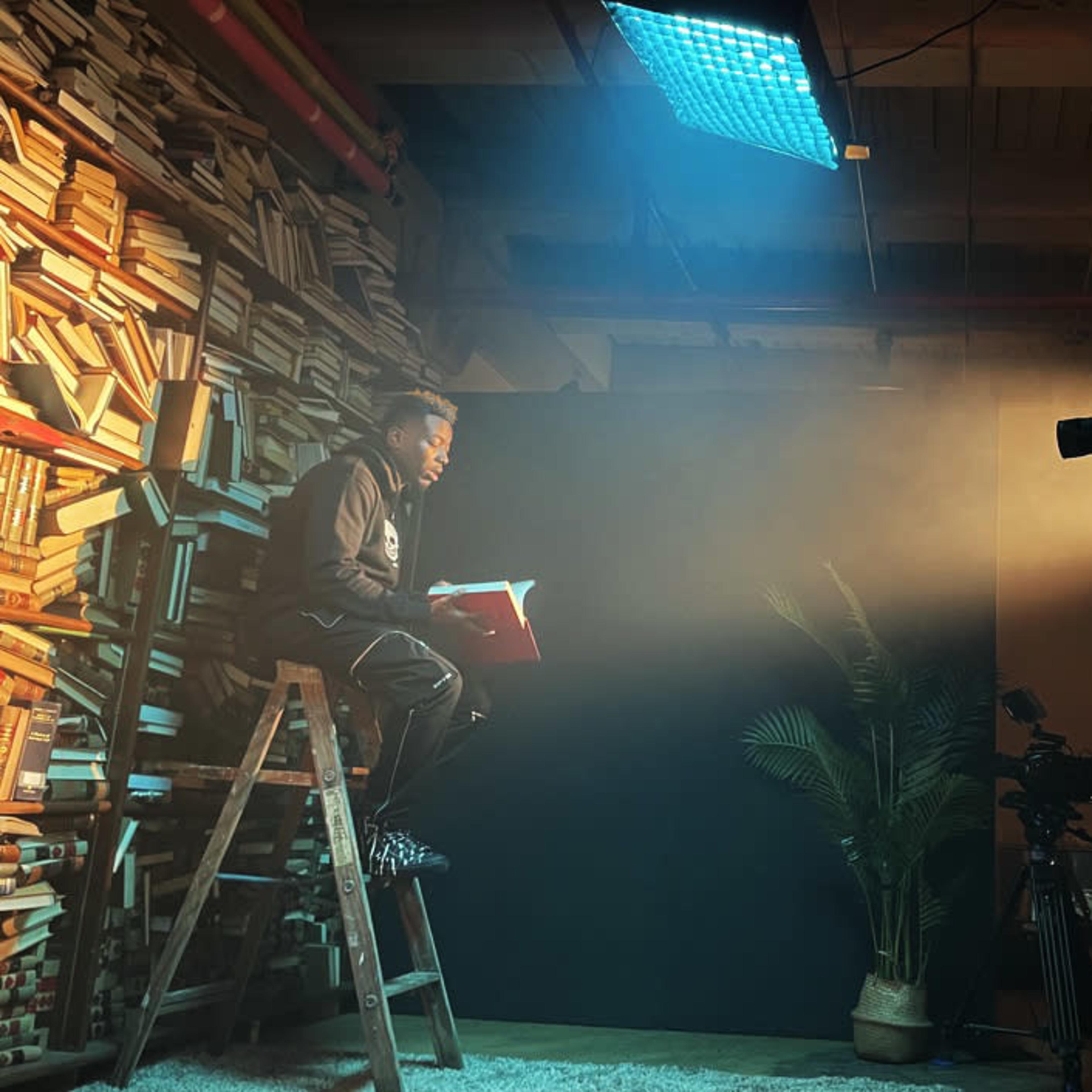 A person sits on a ladder reading a book while another person operates a camera under dramatic lighting in a studio filled with books.