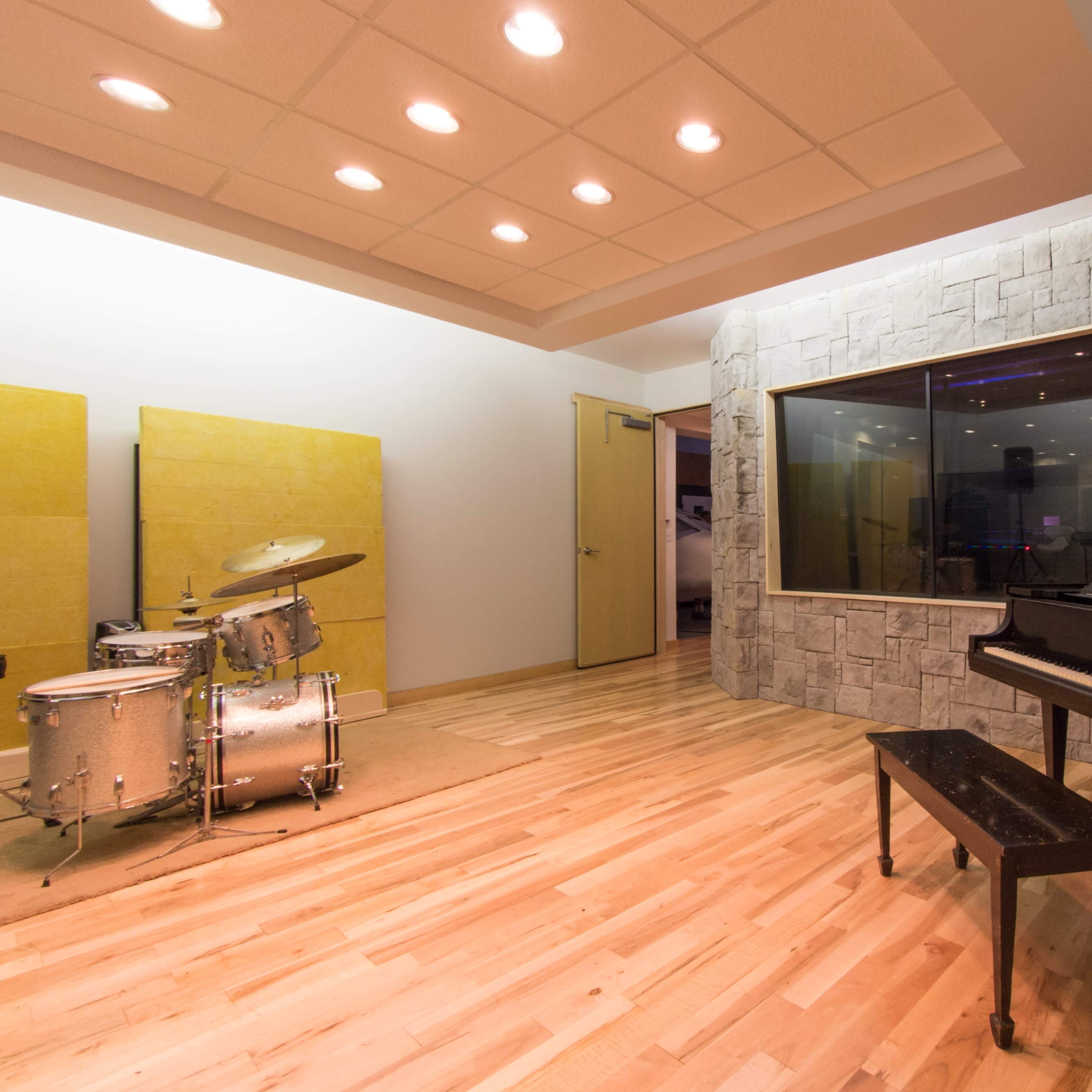 A music studio featuring a drum set, a grand piano, and a glass-enclosed recording booth.