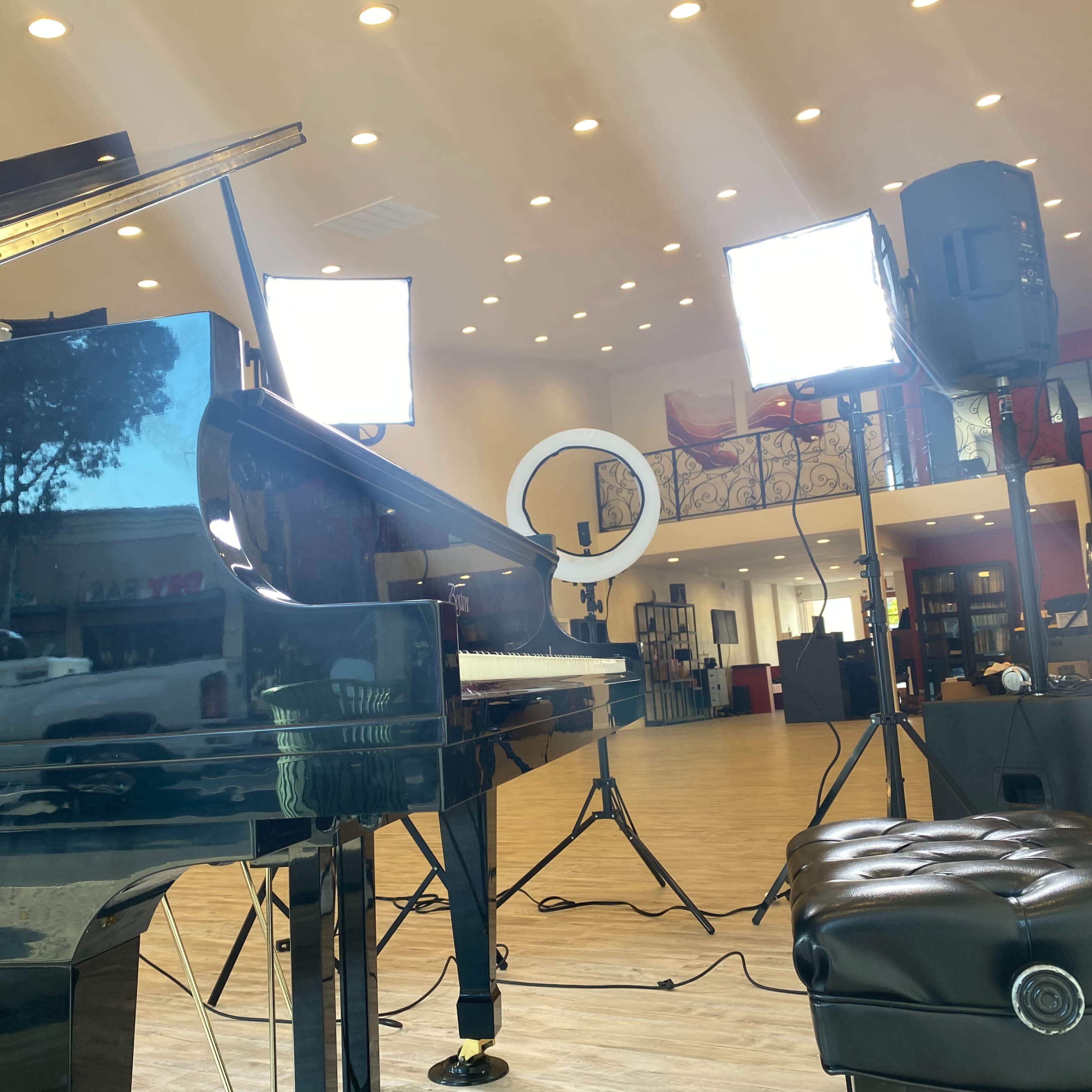A grand piano is positioned in a well-lit room with studio lights and a camera setup, featuring a view of a parked truck outside.