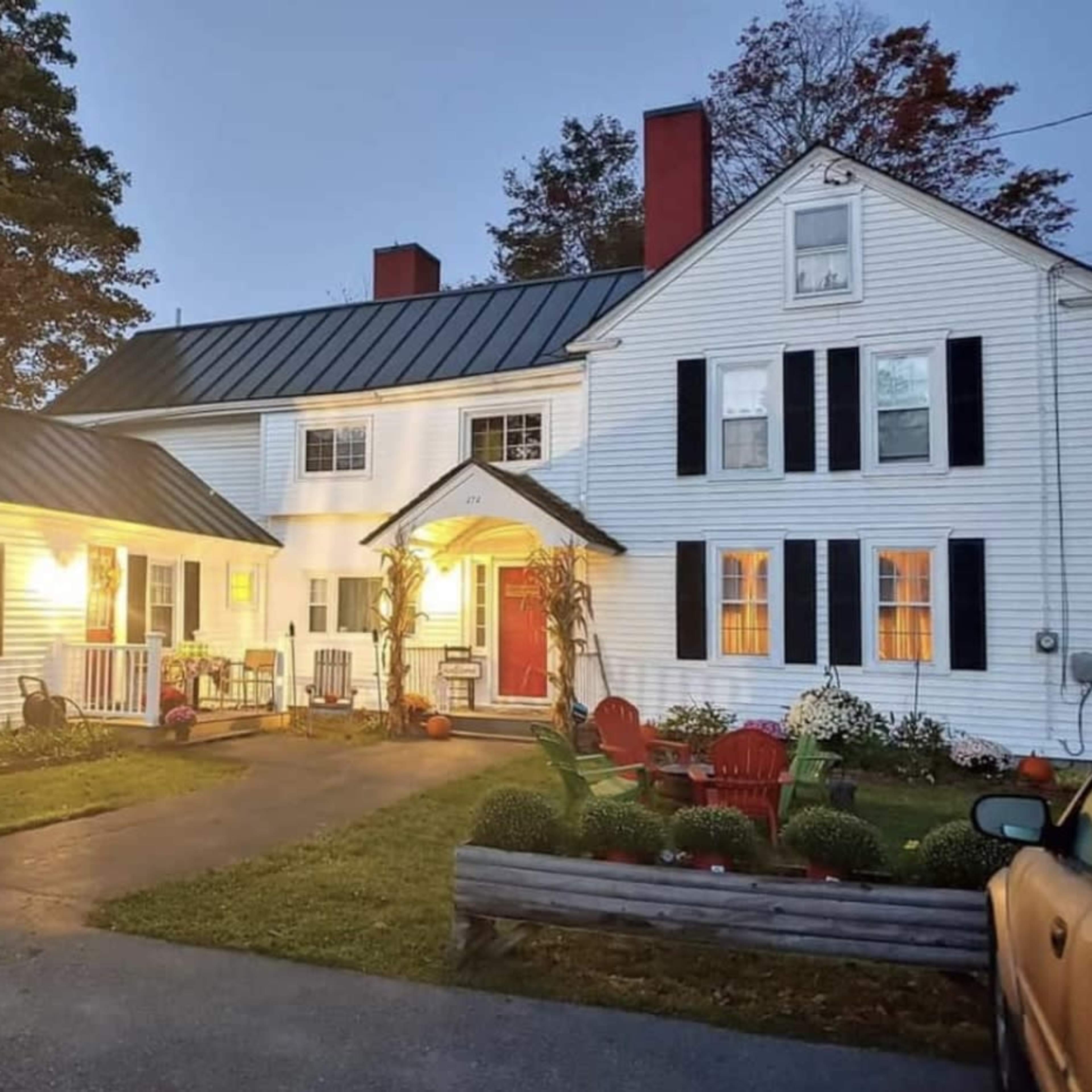 A two-story white house with a black metal roof, a front porch, and a well-maintained yard, illuminated by warm lighting at dusk.