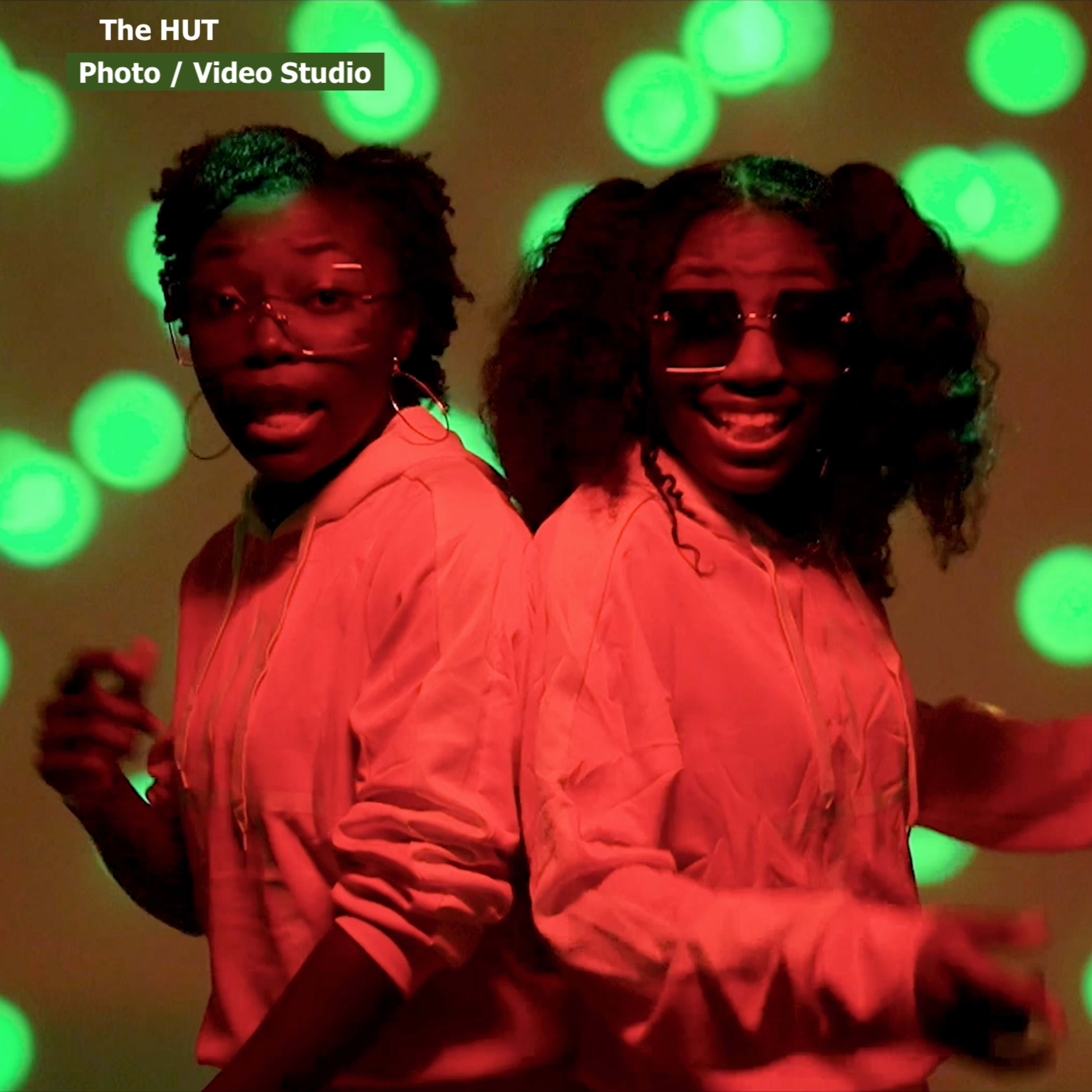 Two individuals stand back to back in a photo studio, wearing hoodies and sunglasses against a green bokeh background.