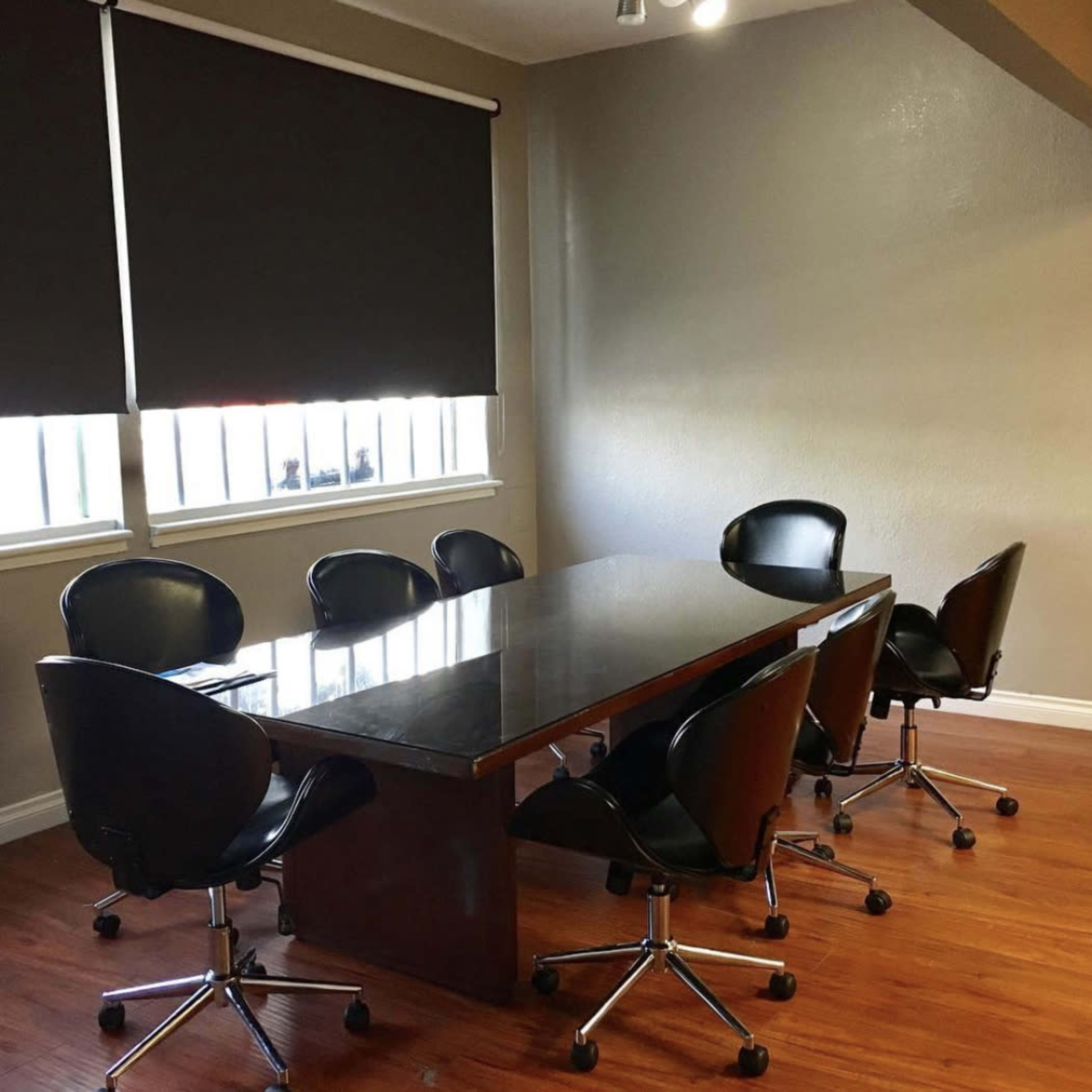 A conference room features a long wooden table surrounded by six black rolling chairs, with two large black blinds covering the windows.
