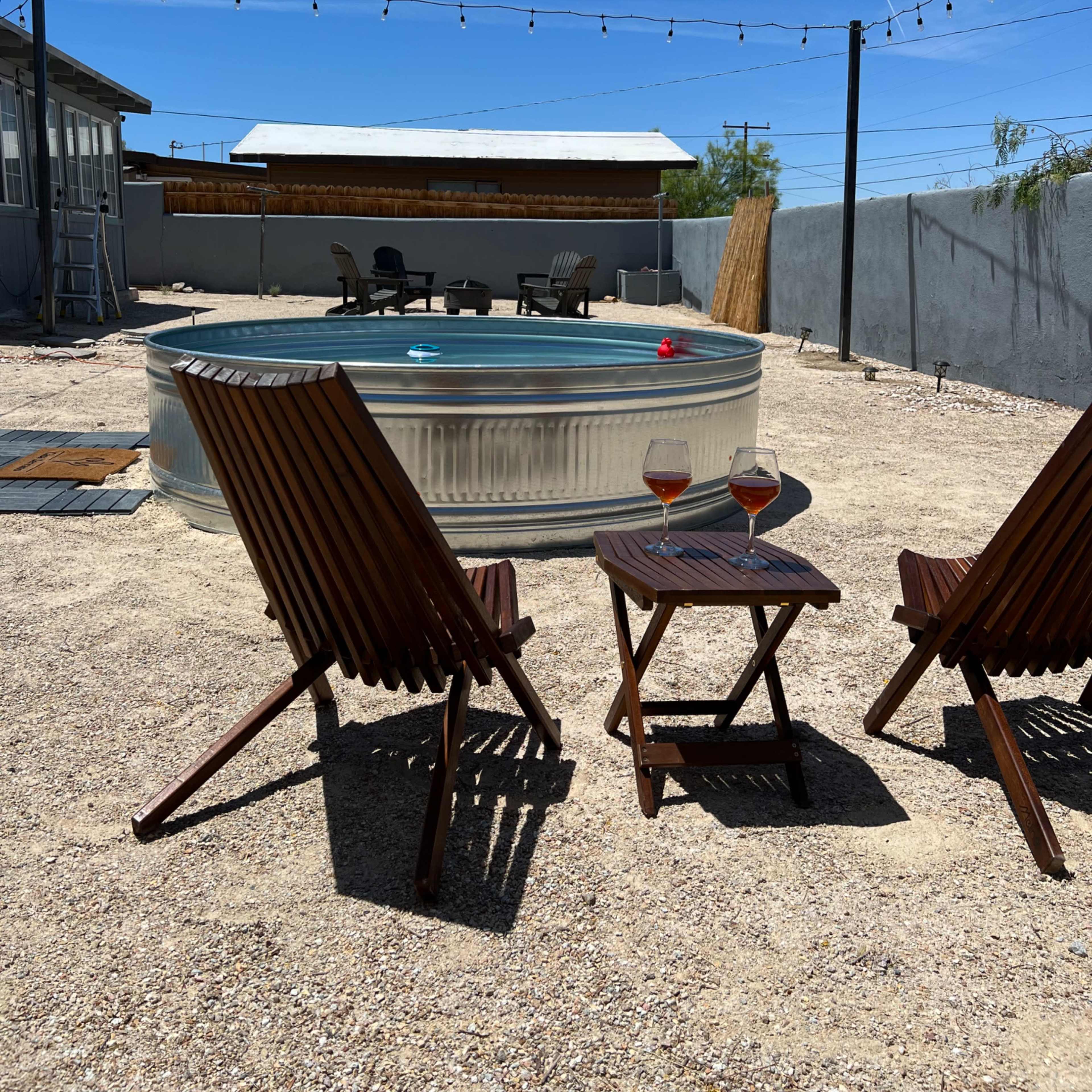 A circular pool sits in a gravel backyard with two wooden lounge chairs and a small table holding two glasses.