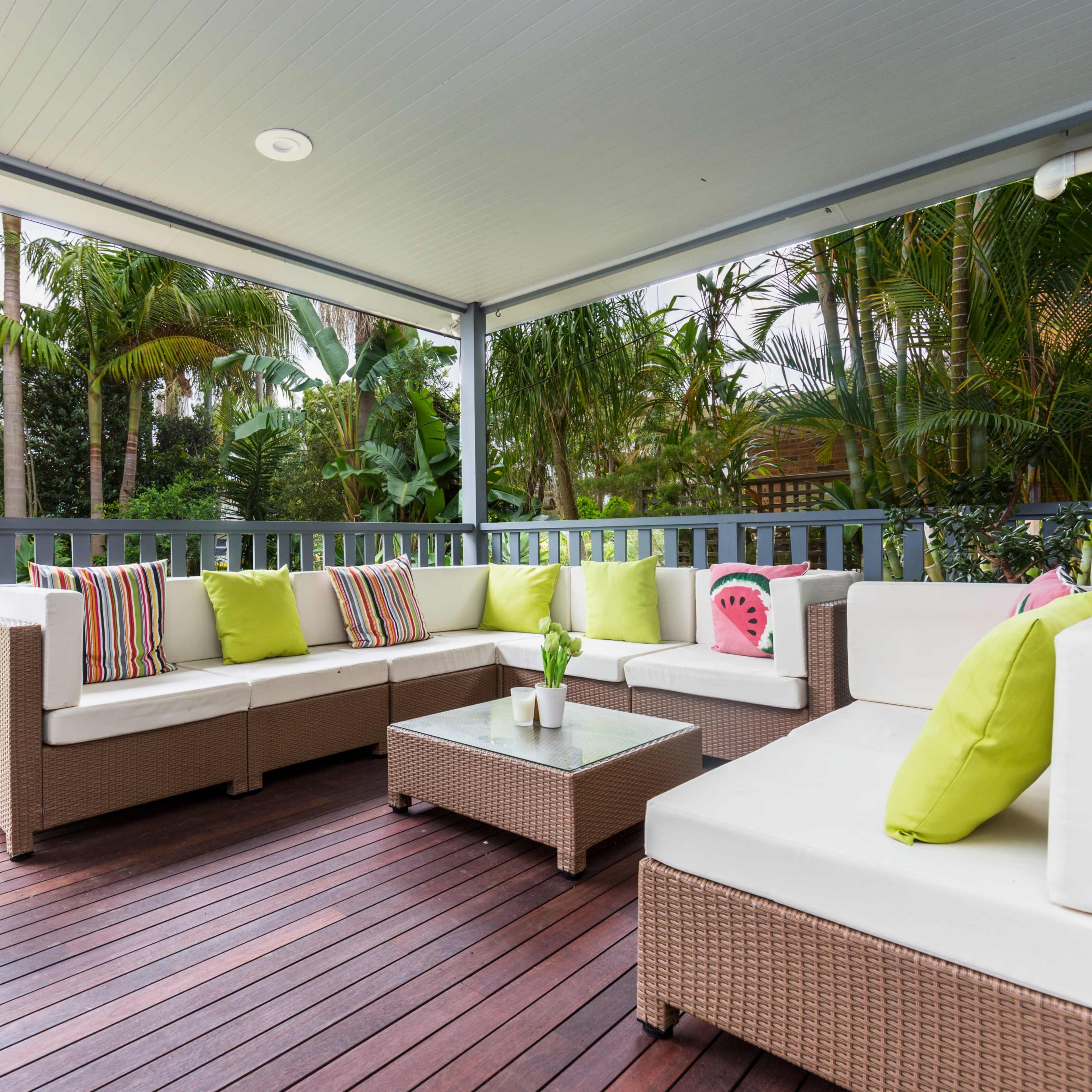 A covered porch features a sectional sofa with brightly colored cushions and a coffee table surrounded by greenery.