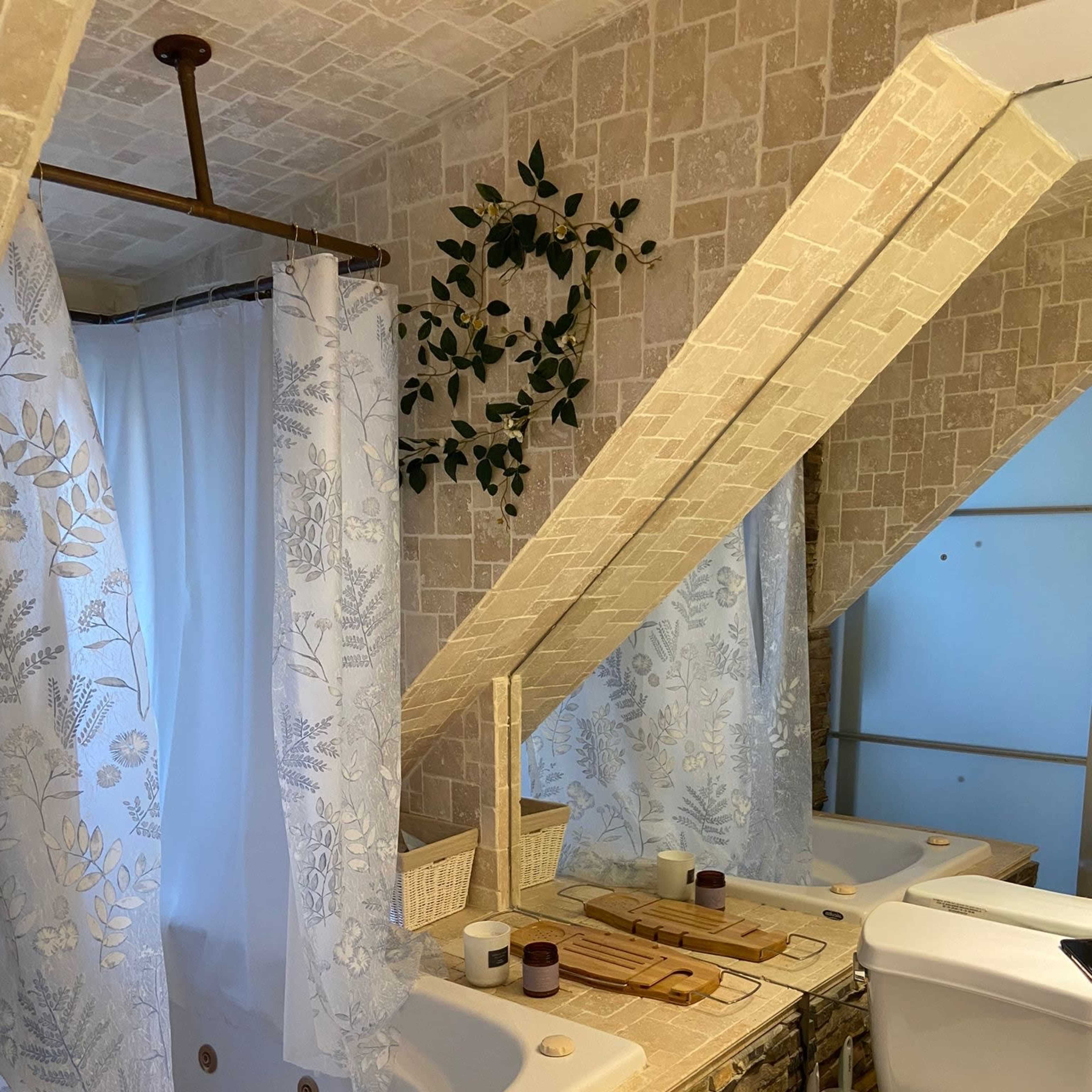The bathroom features a corner jetted tub with a shower curtain, a double vanity with wooden accessories, and a toilet, all set against a backdrop of stone and tile.