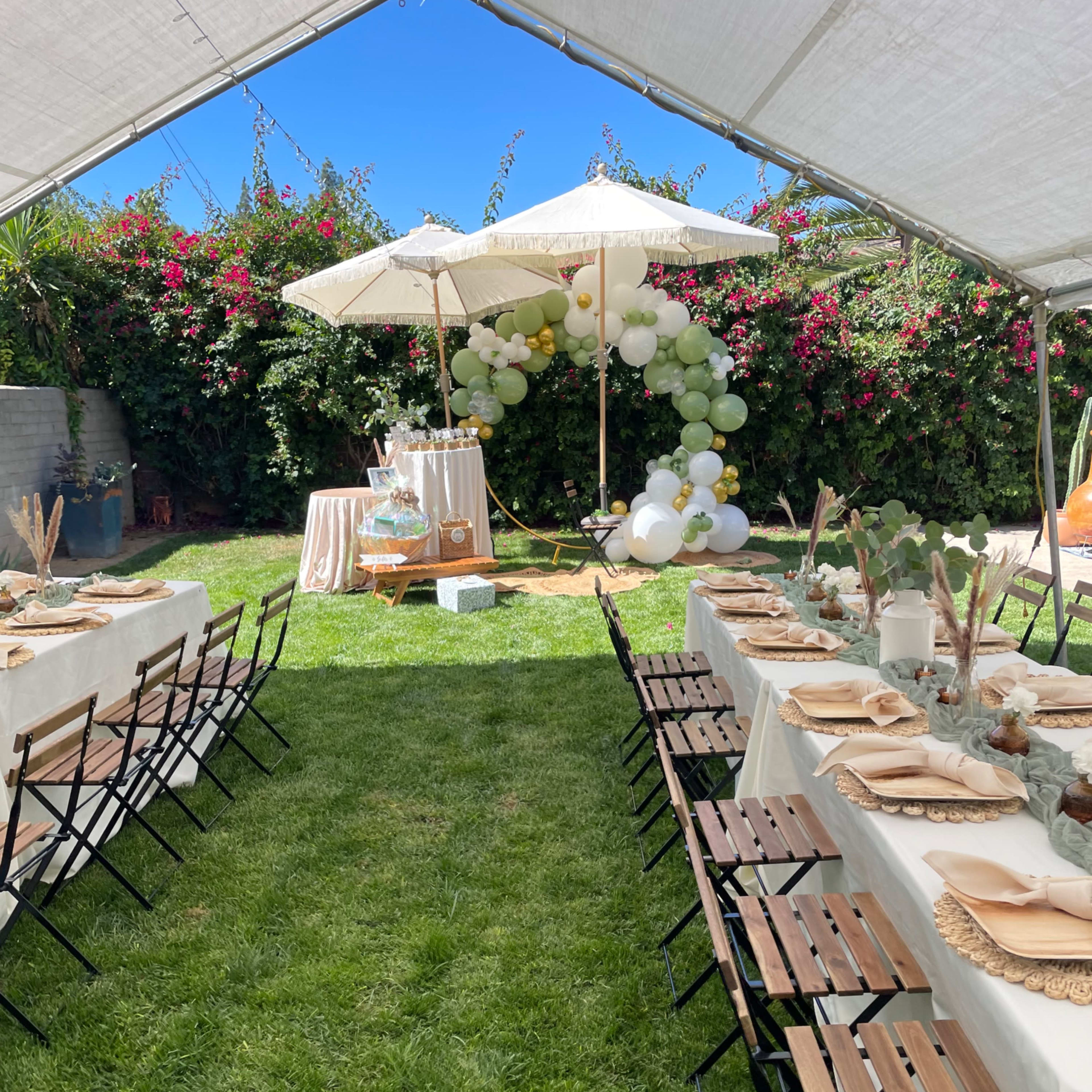 A tented outdoor setting features long tables with beige tablecloths, folding chairs, and decorative umbrellas, surrounded by greenery and floral arrangements.