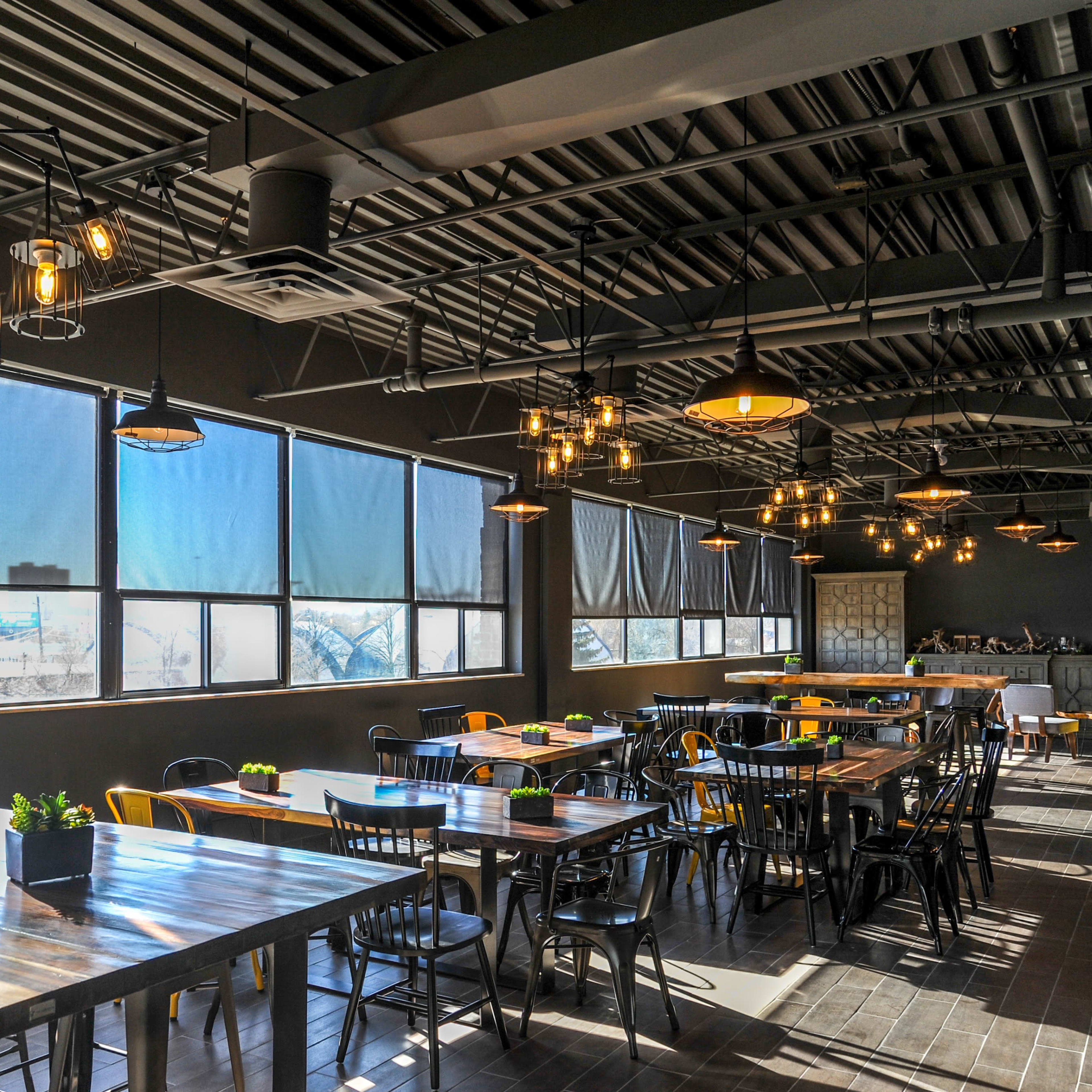 A spacious restaurant interior features multiple wooden tables and chairs under industrial-style lighting, with large windows allowing natural light to illuminate the room.