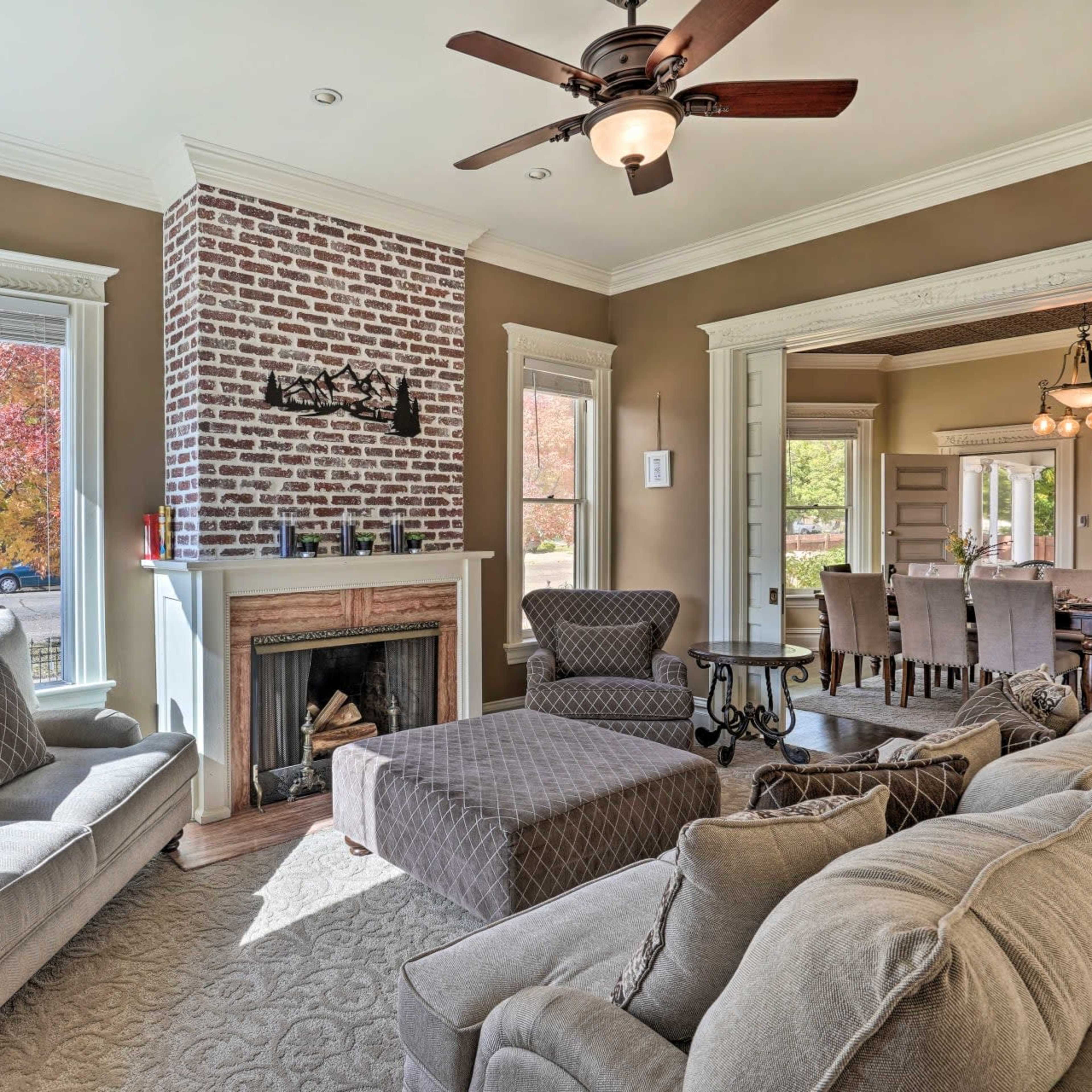 A cozy living room features a brick fireplace, large windows, and comfortable seating with a view of an outdoor scene.