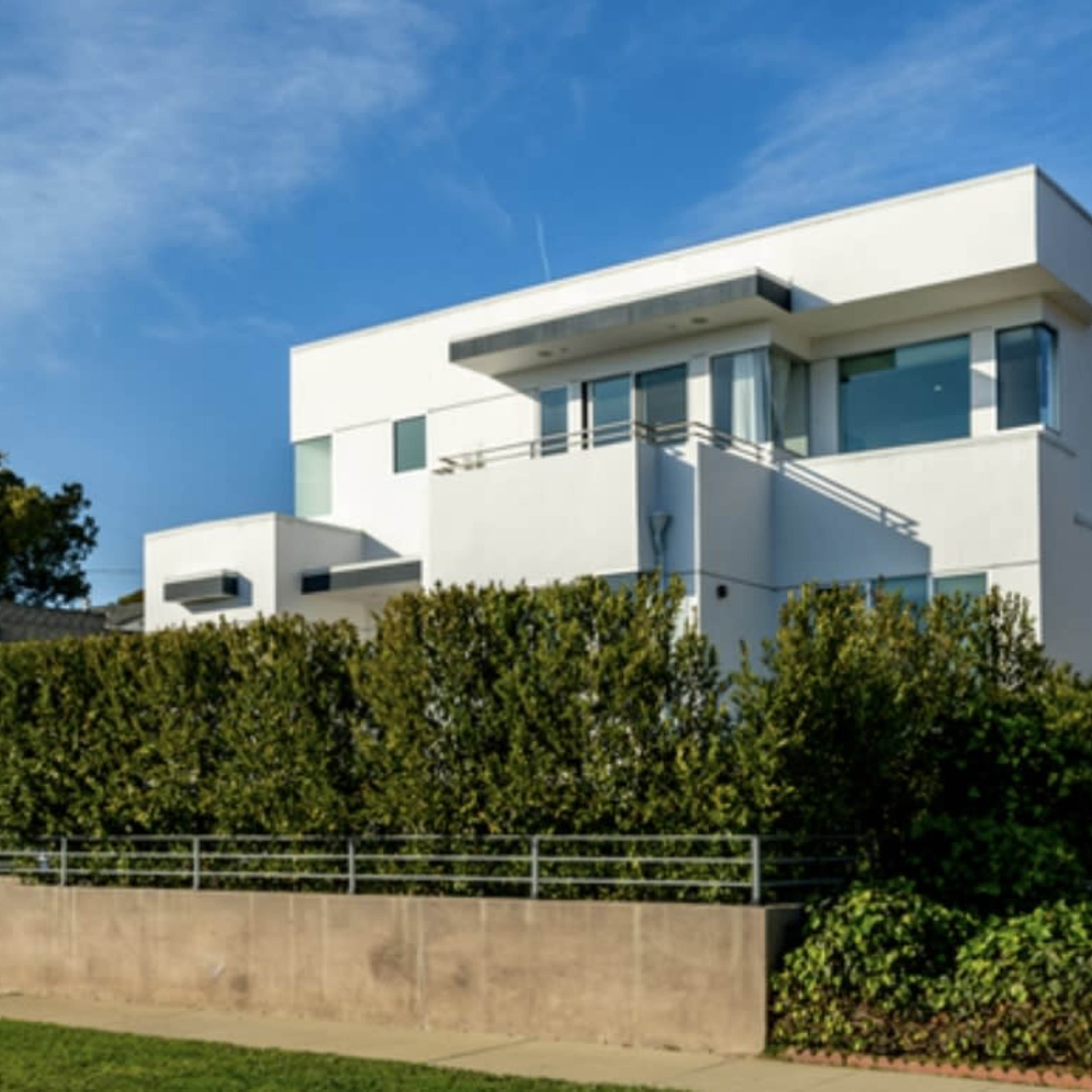 A modern, two-story white house is set behind a tall hedge with a concrete wall along the front.