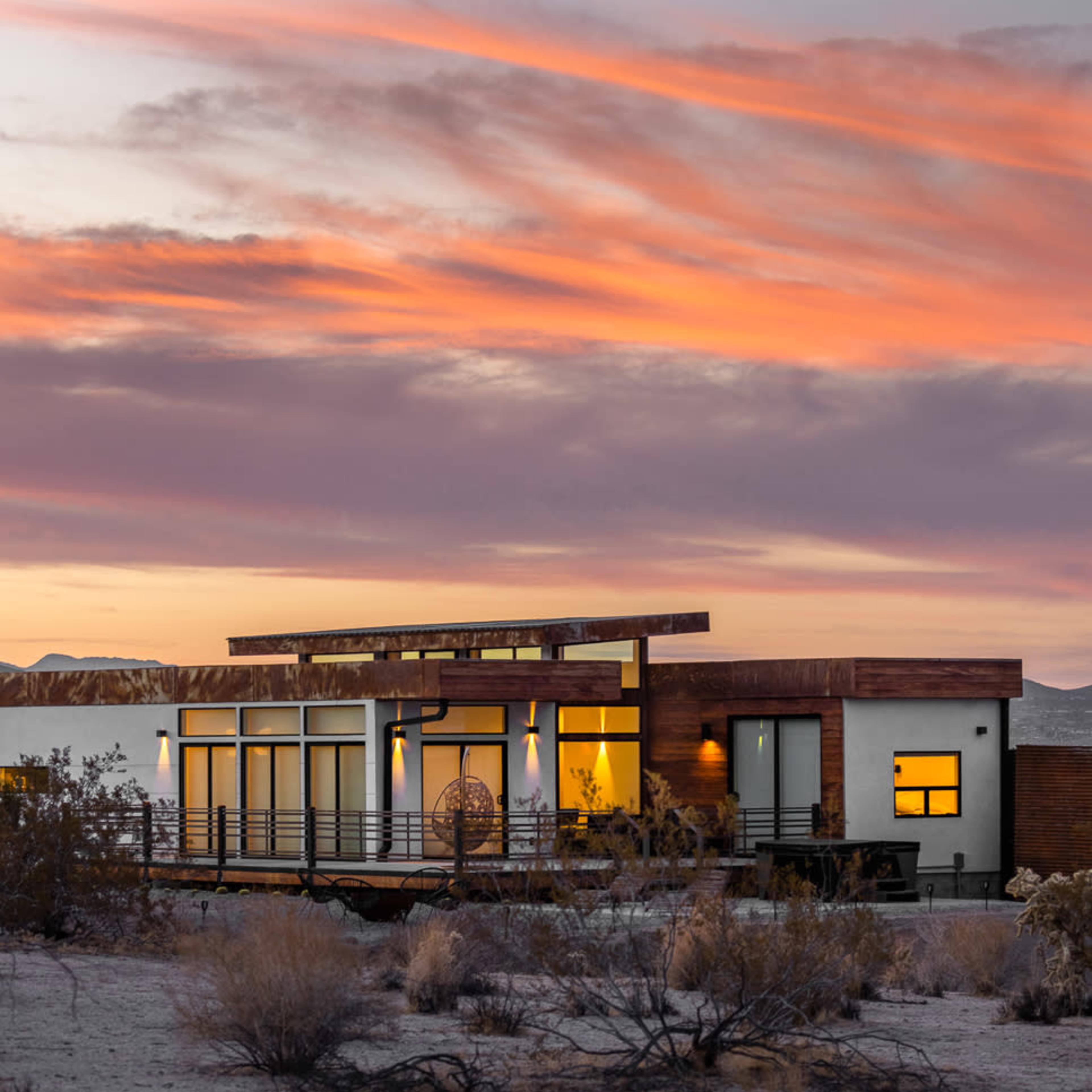 A modern house with large windows and a wooden exterior is set against a colorful sunset sky over a desert landscape.