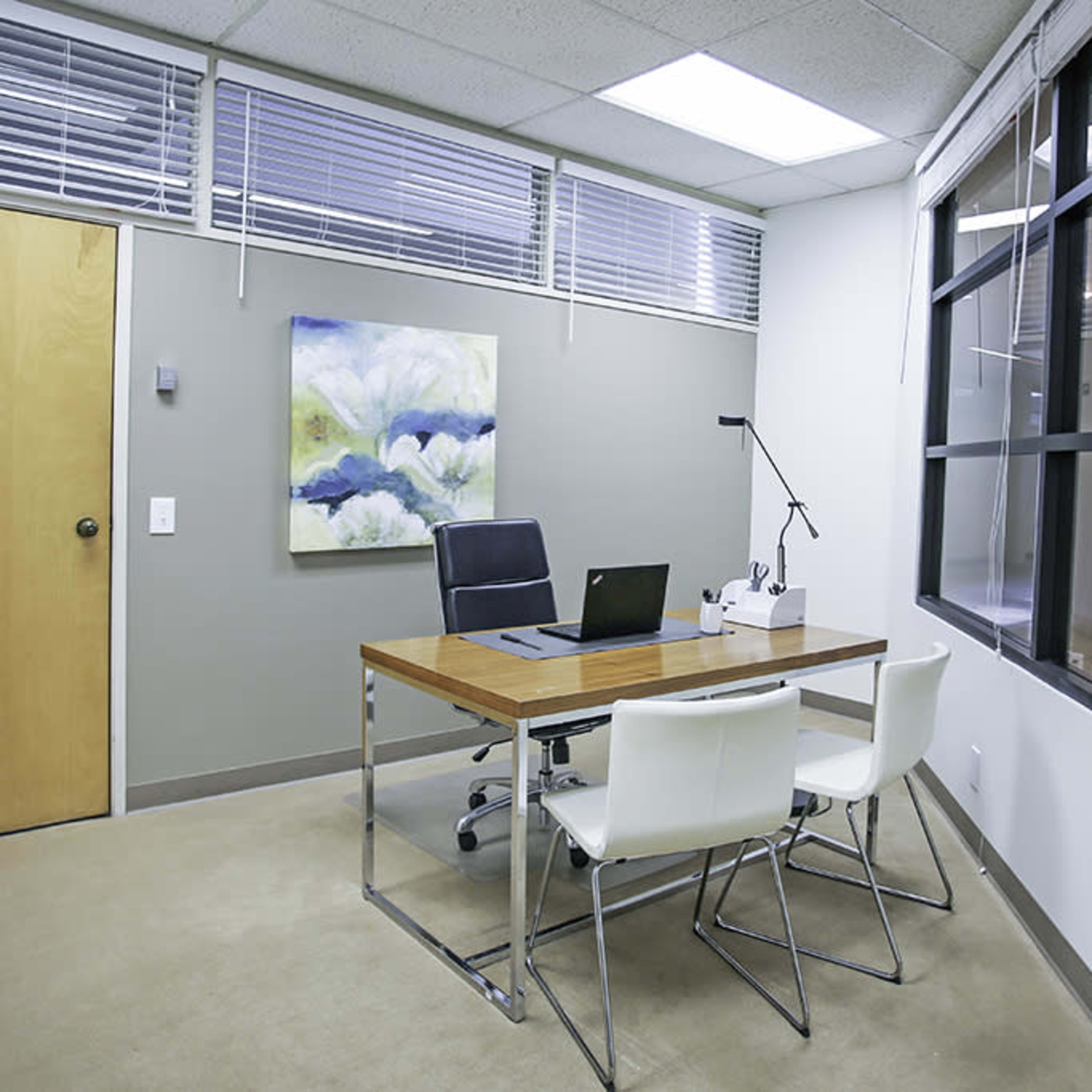 A simple office space with a wooden desk, a black chair, two white chairs, and a large window letting in natural light.