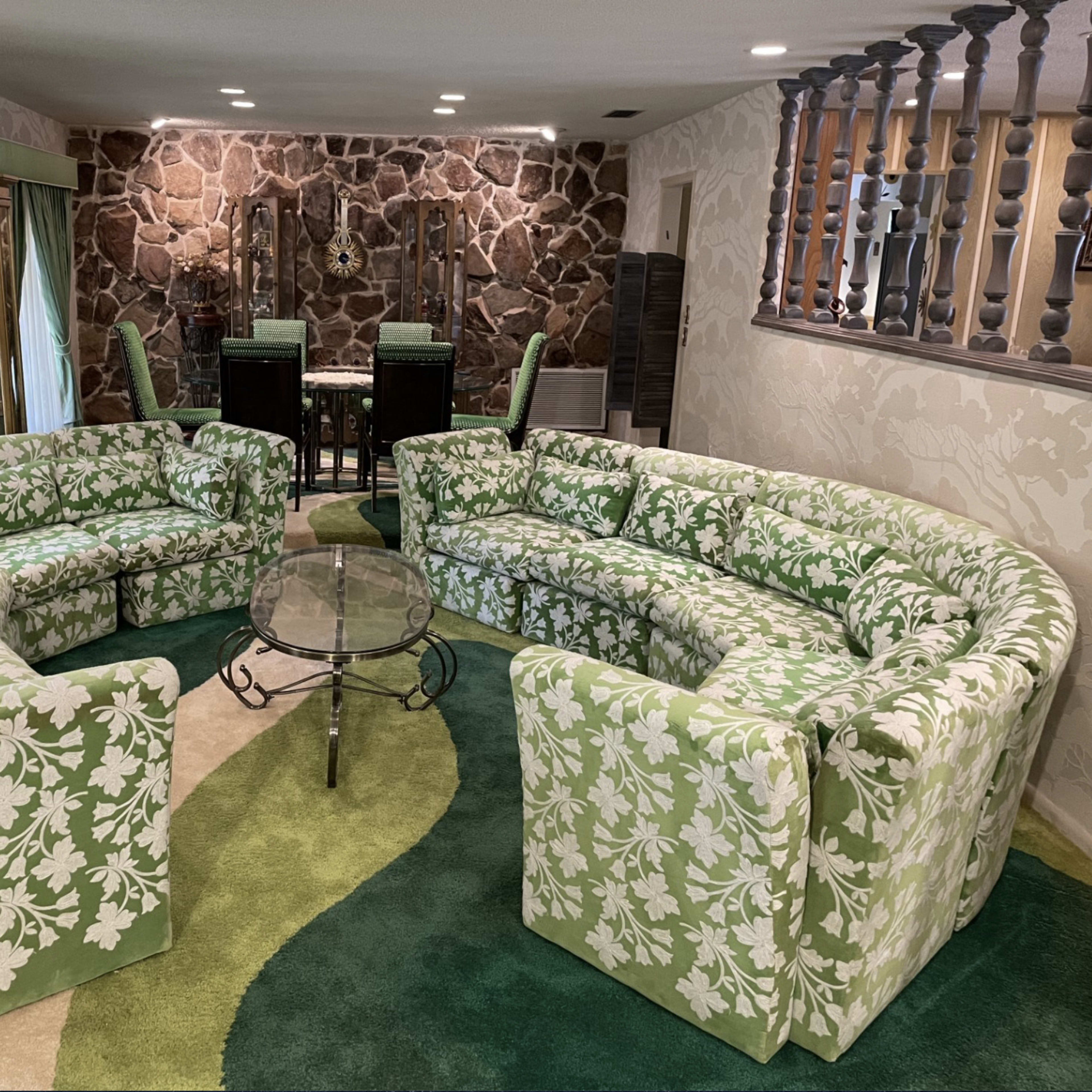 The scene features a circular arrangement of green floral-patterned sofas surrounding a glass coffee table on a green carpet in a room with stone walls and a dining area in the background.