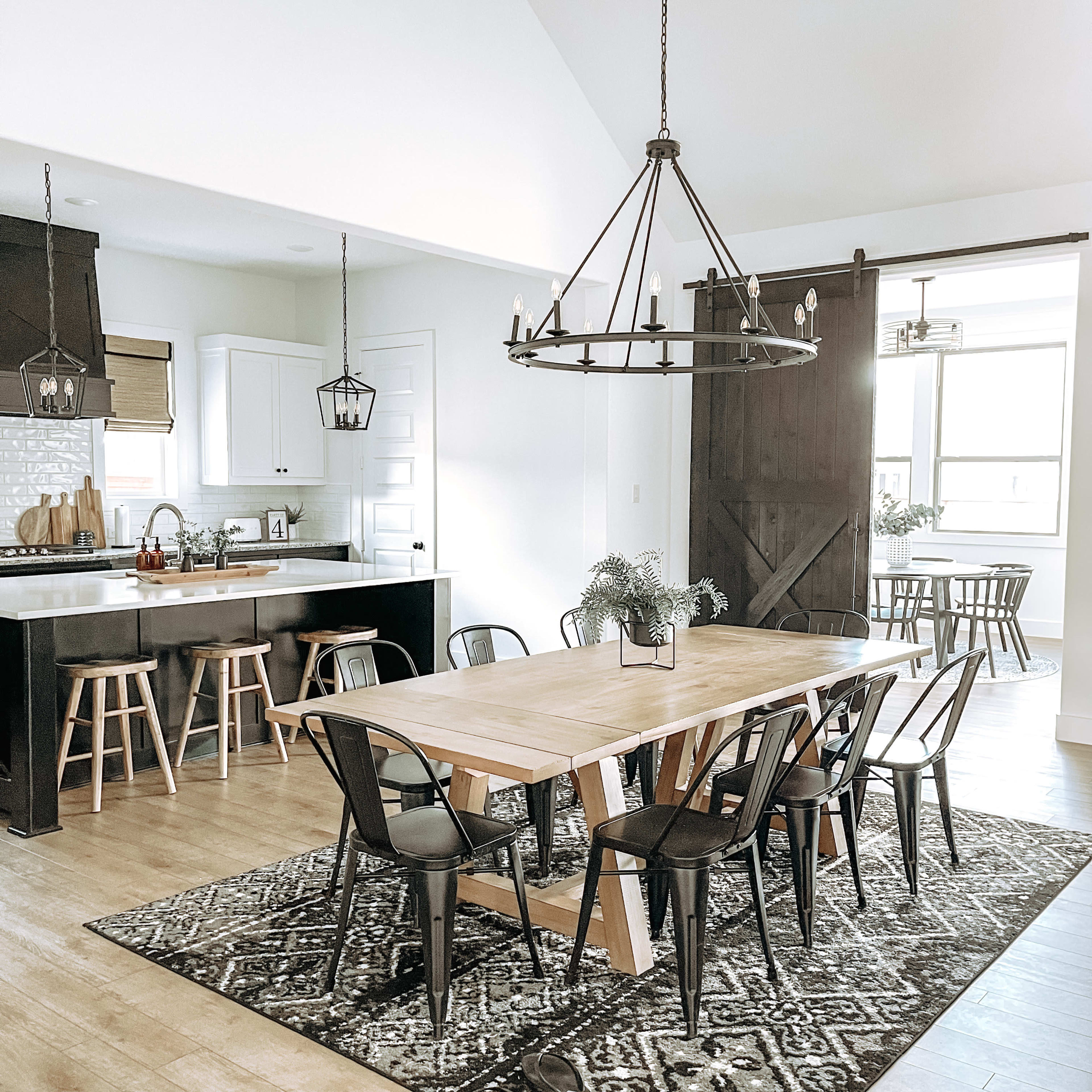 A modern dining area with a wooden table surrounded by black chairs, a light fixture overhead, and a view of a kitchen area in the background.