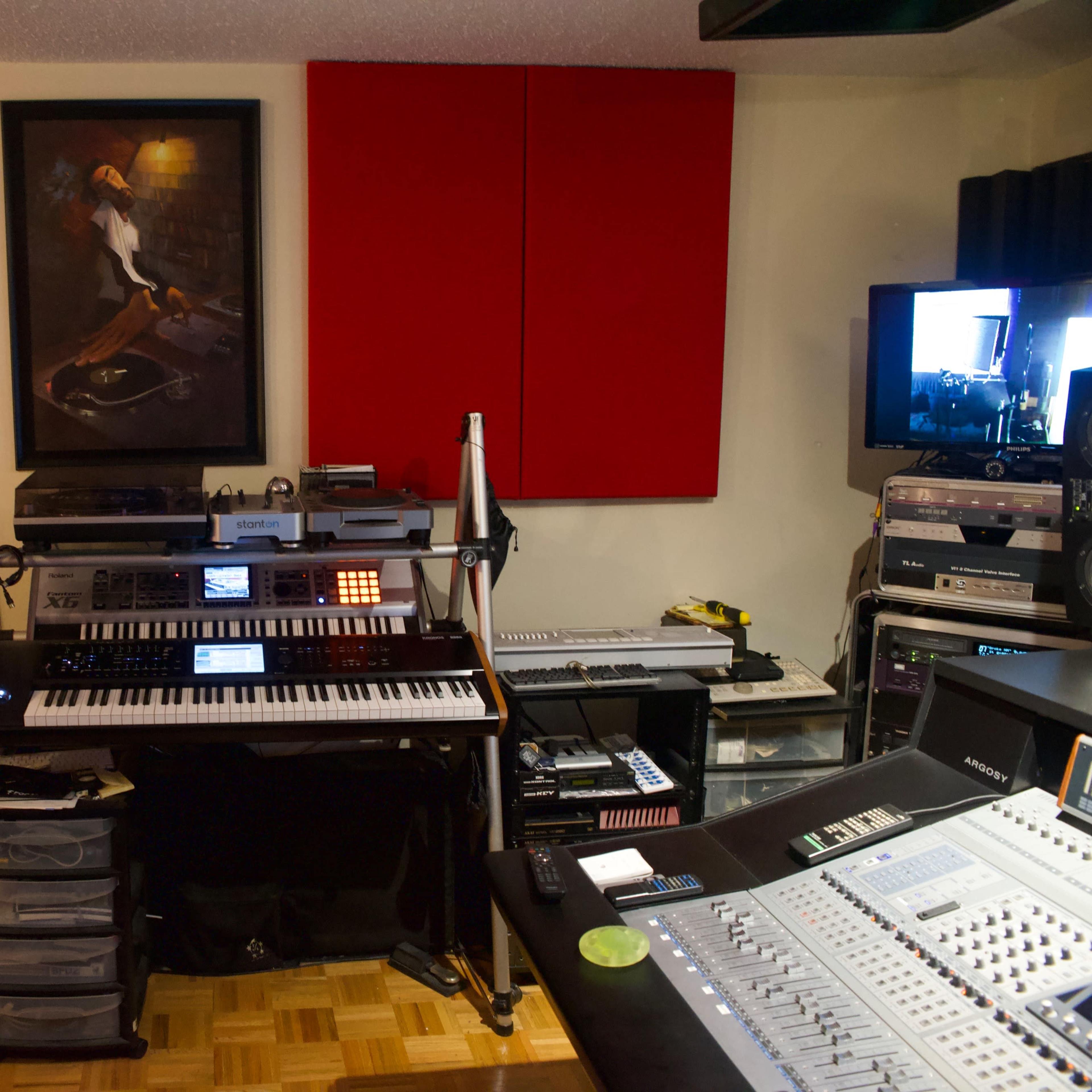 A music production studio featuring an array of equipment, including a keyboard, mixing console, and wall-mounted screens, with red sound panels and artwork on the walls.