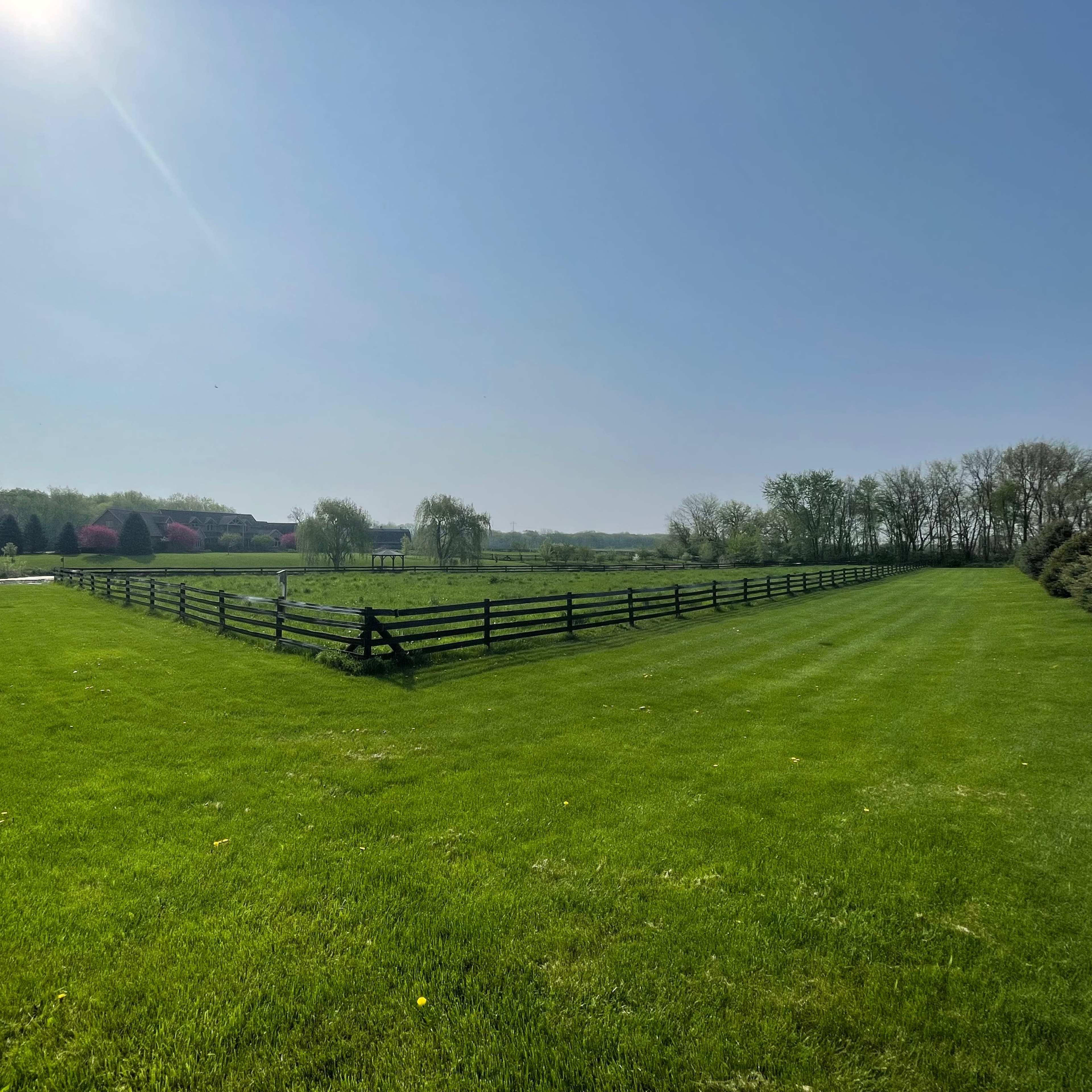 A large green field bordered by a black wooden fence under a clear blue sky.