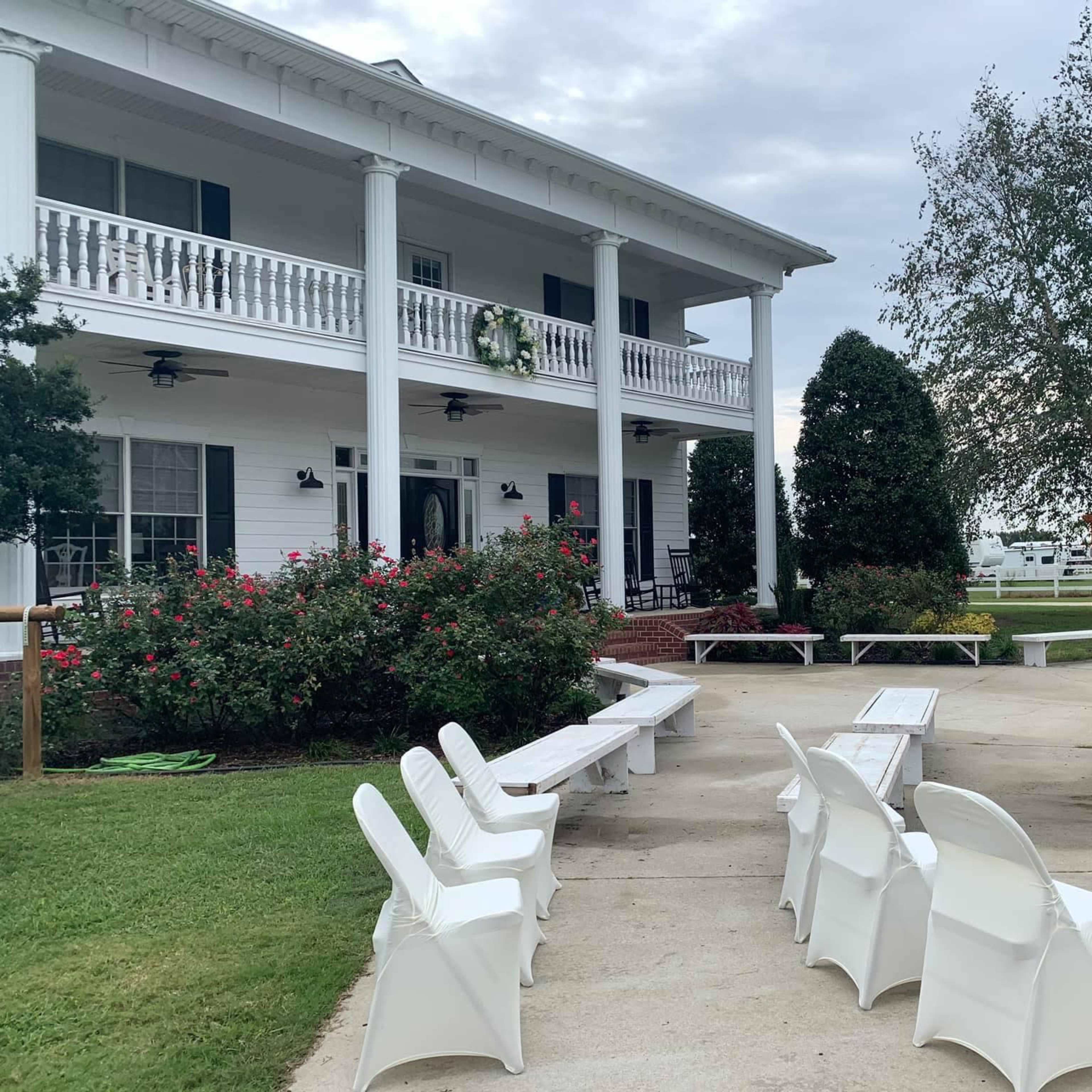 A white house with a large porch and multiple columns is surrounded by green grass, benches, and a swing set in the yard.