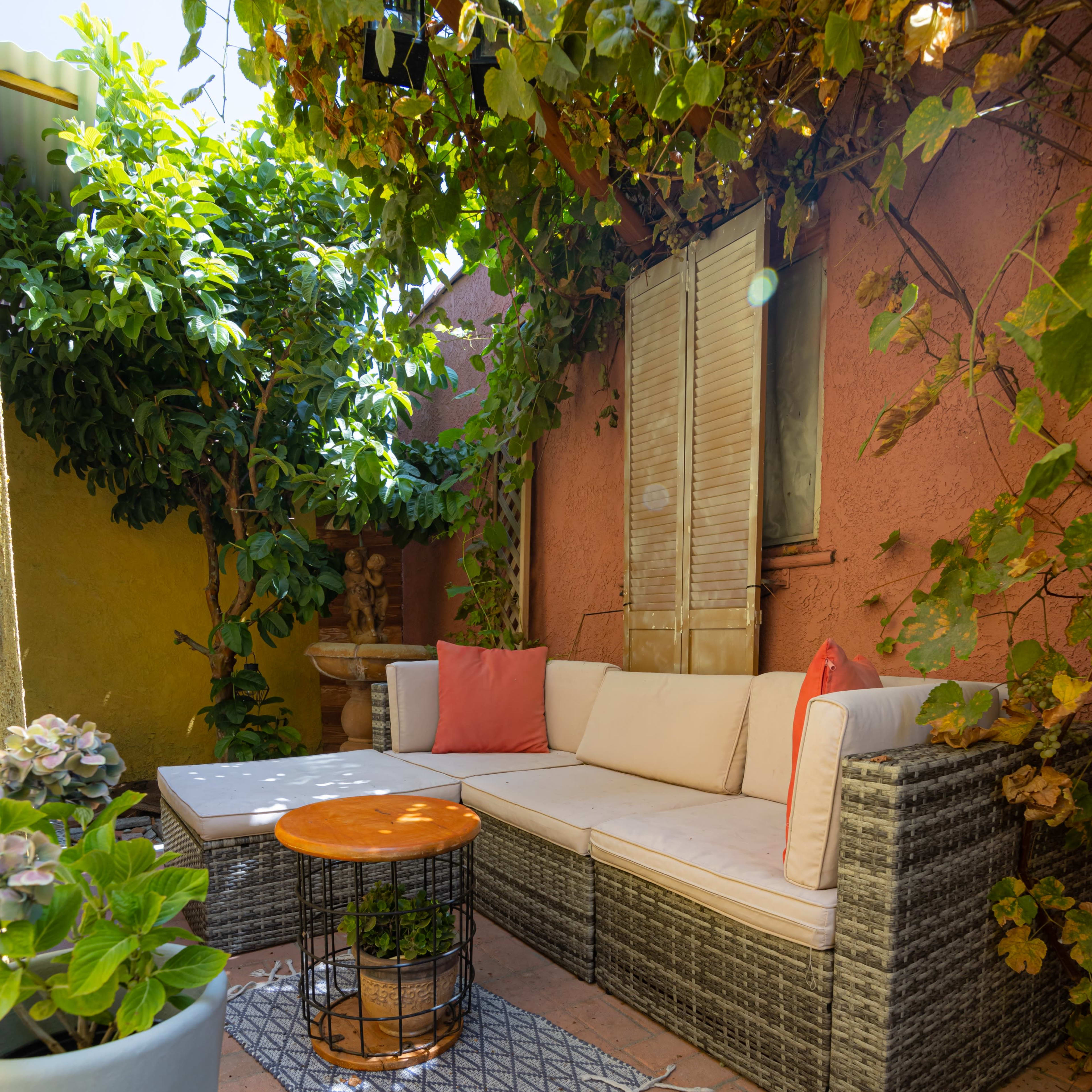 A cozy outdoor seating area features a wicker sofa with cushions, a wooden coffee table, and surrounding greenery against a colorful wall.