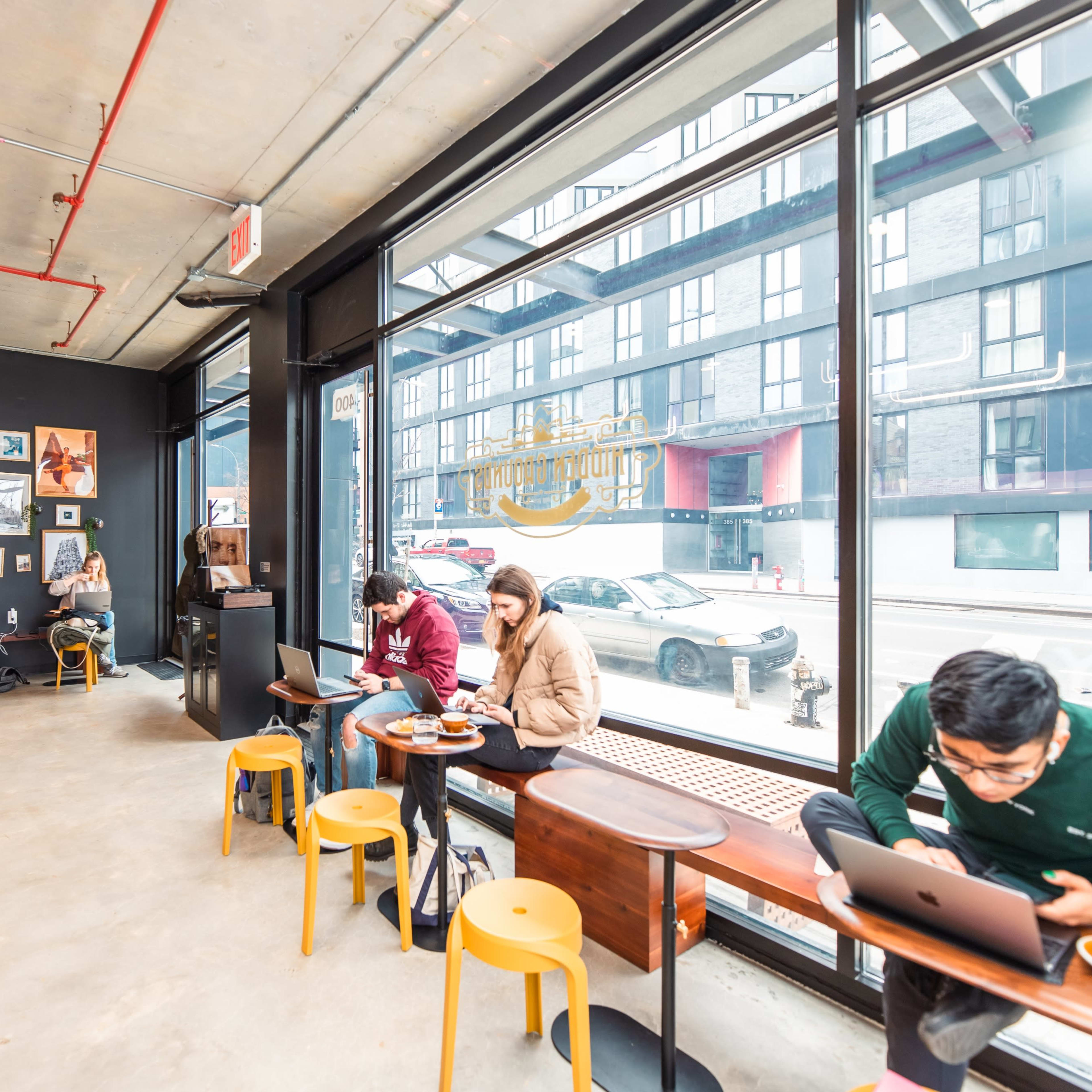 A cozy café interior features patrons seated at tables, some working on laptops, while large windows offer a view of the street outside.