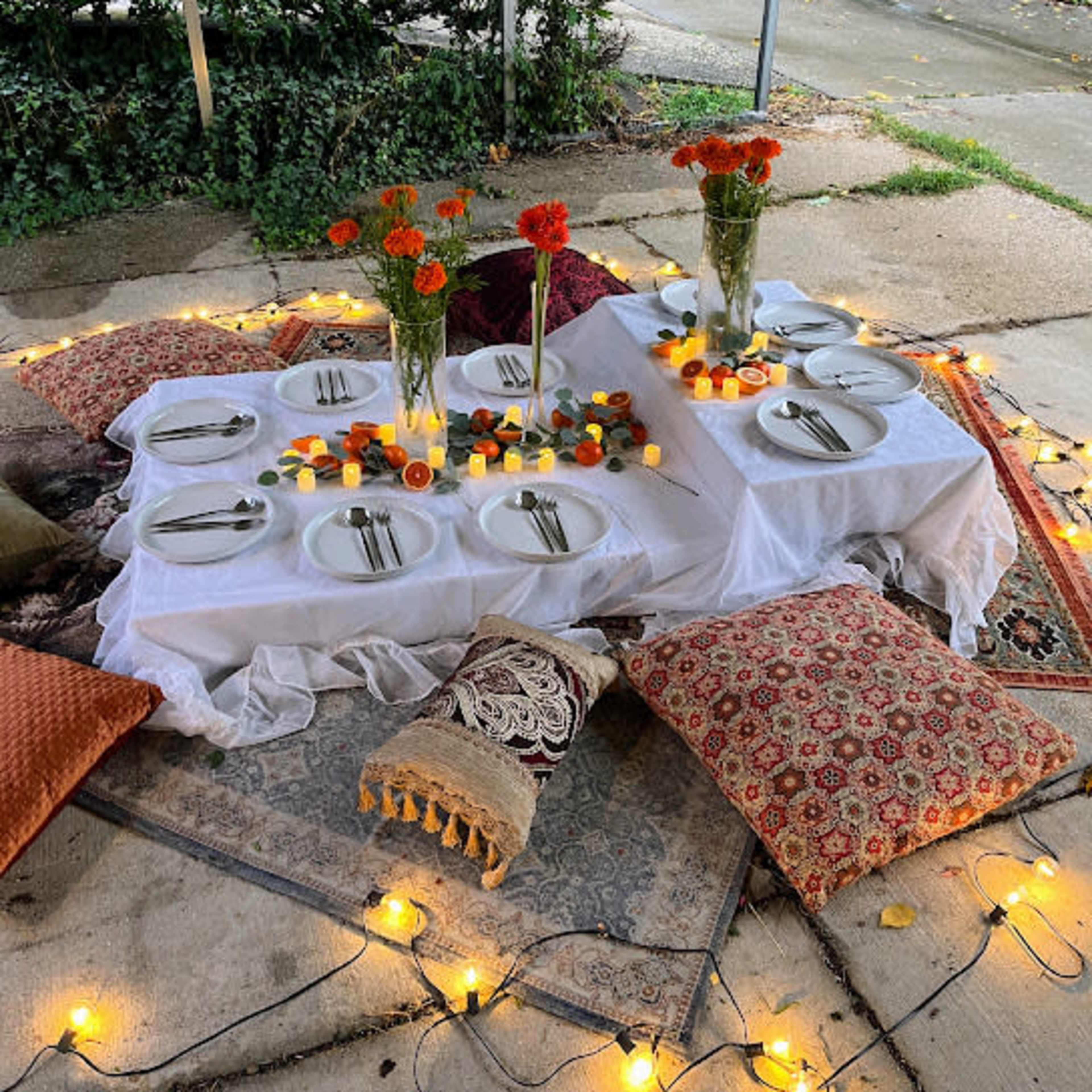 A picnic setup with a white tablecloth laid on the ground, surrounded by plates, cutlery, and decorated with flowers and candles, under a covered area.