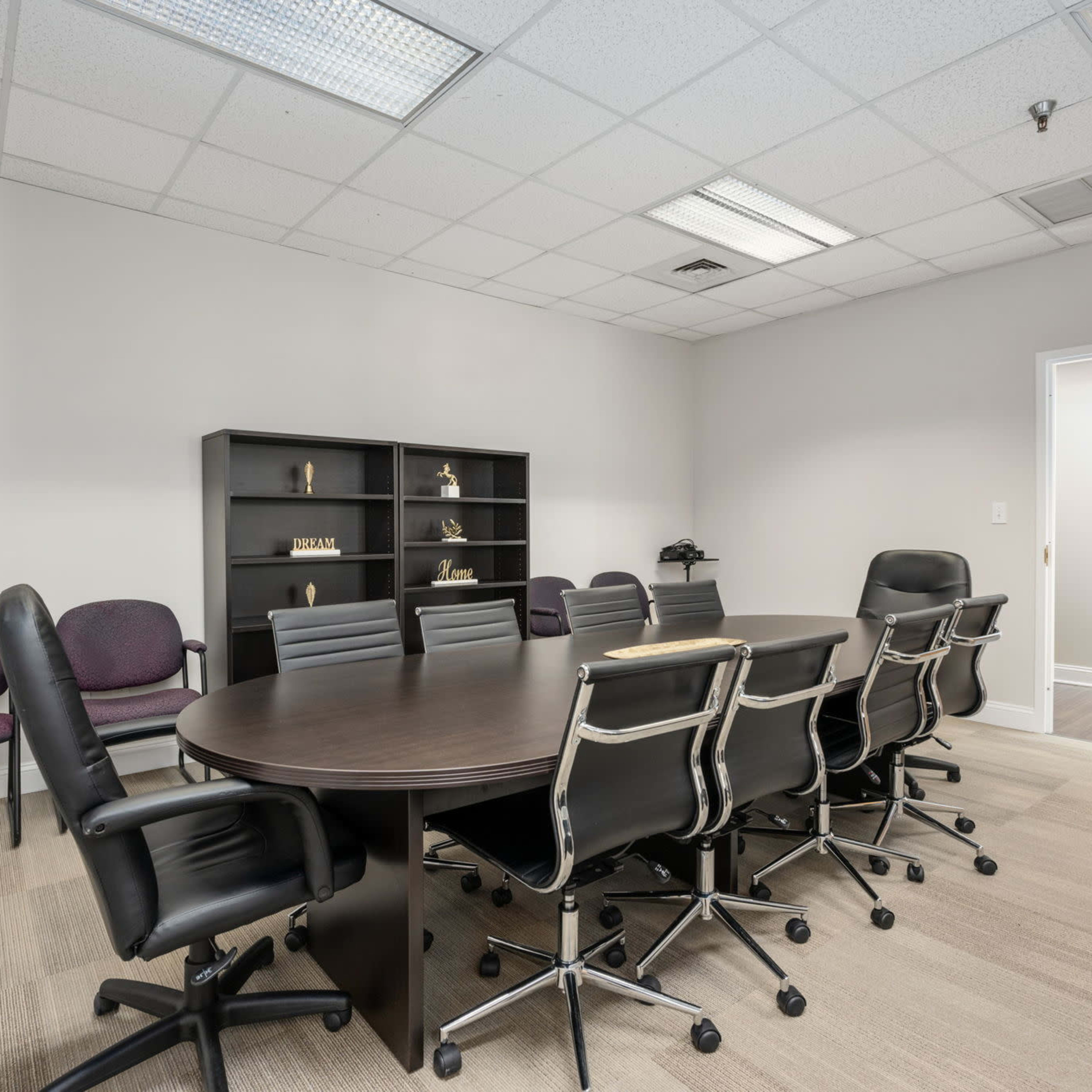 A conference room features a large oval table surrounded by black leather chairs, with a shelving unit against the wall and several empty chairs at one side.