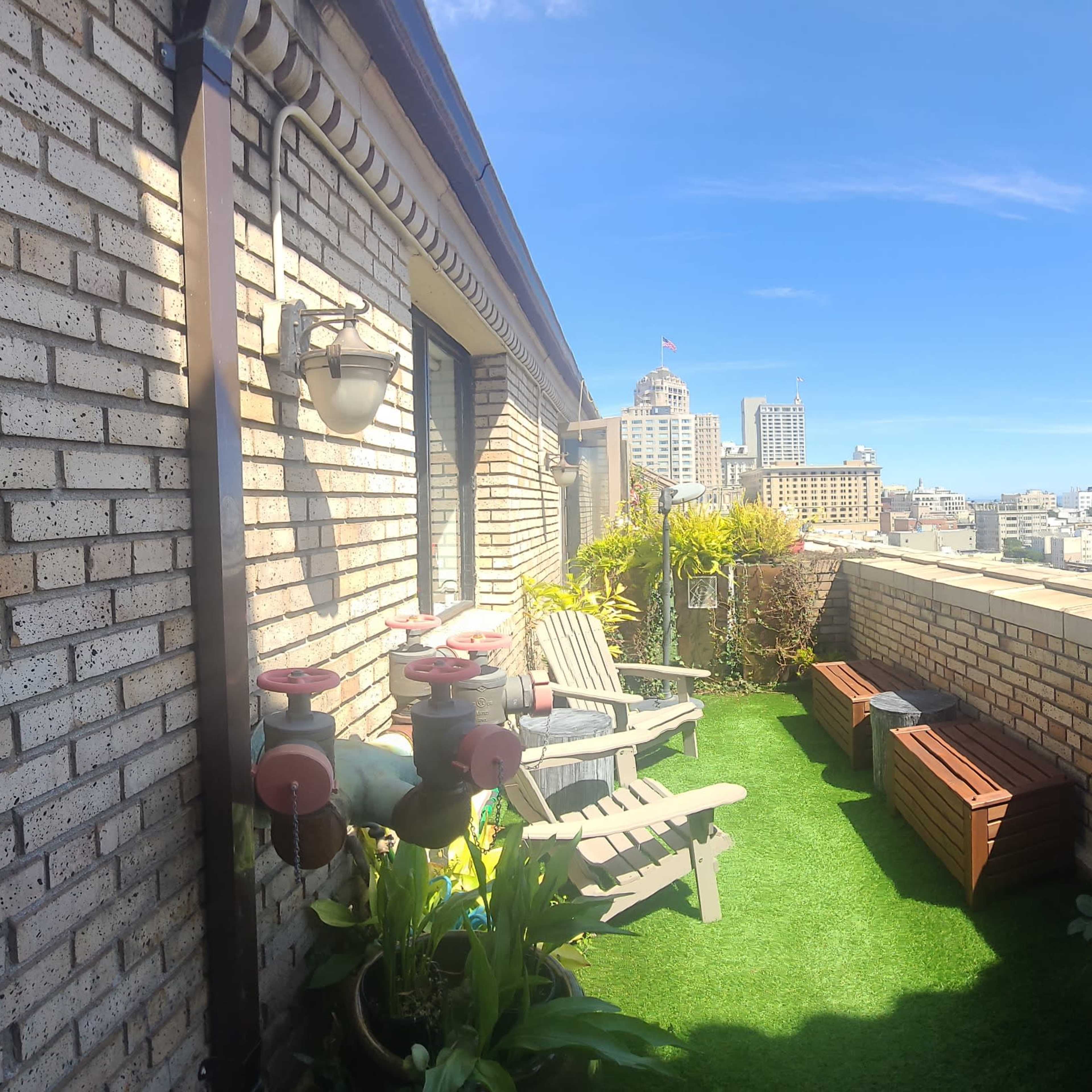 A rooftop terrace with green artificial grass, potted plants, and outdoor seating, overlooking a city skyline.