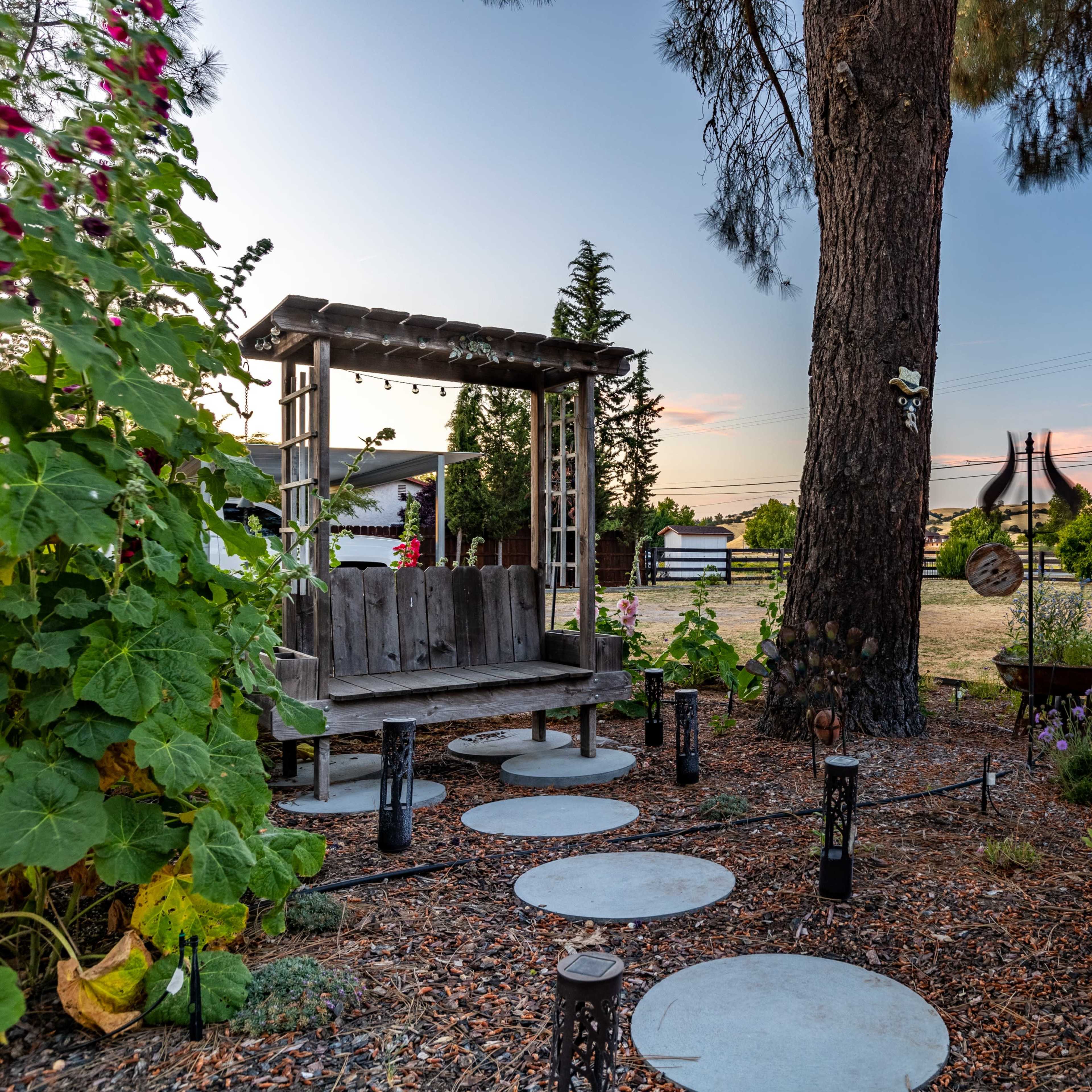 A wooden bench sits under a tree, surrounded by stepping stones and landscaping, with a grassy field and a fence in the background.