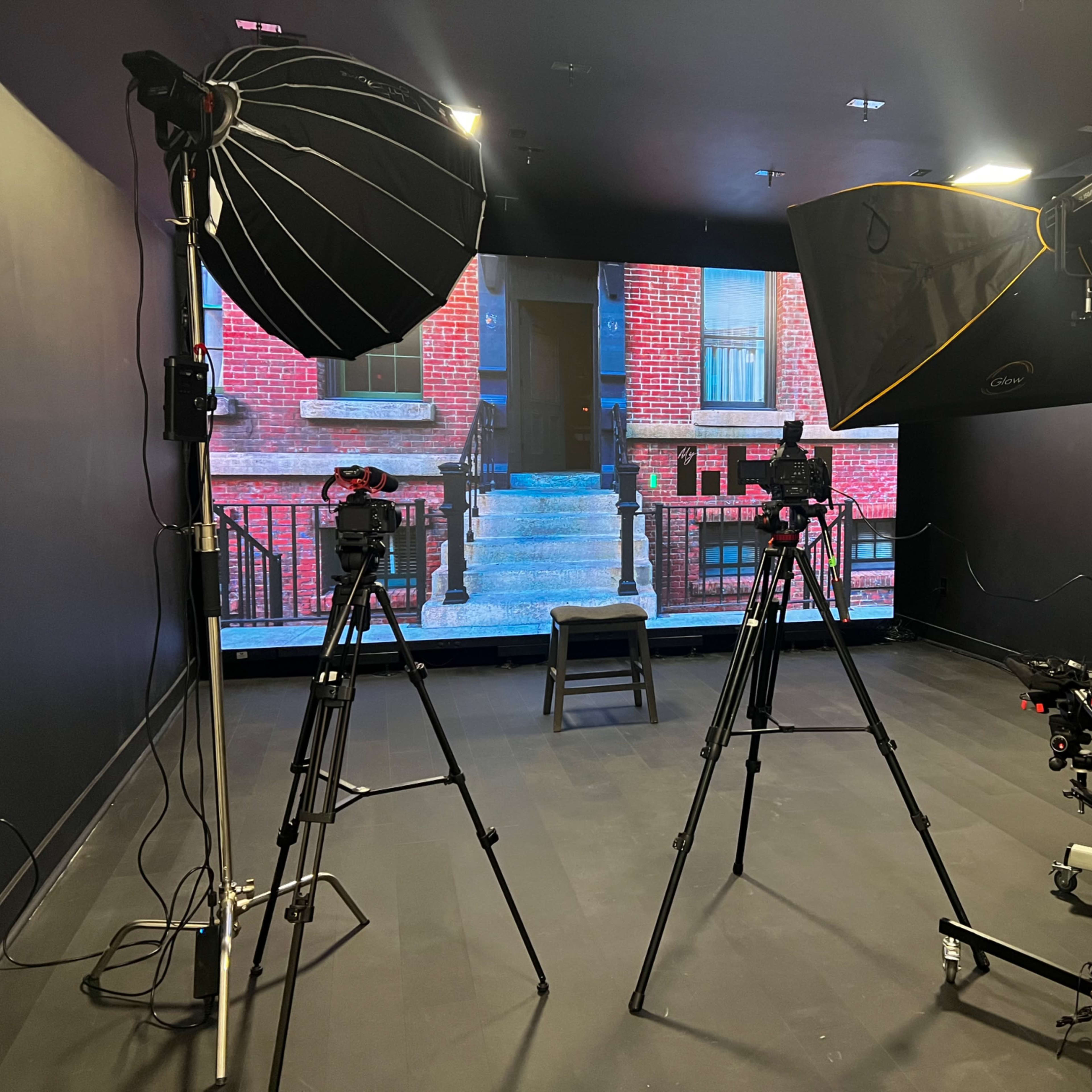 A photography setup with two large softbox lights, cameras on tripods, and a projector displaying a brick building on a screen in a dark room.