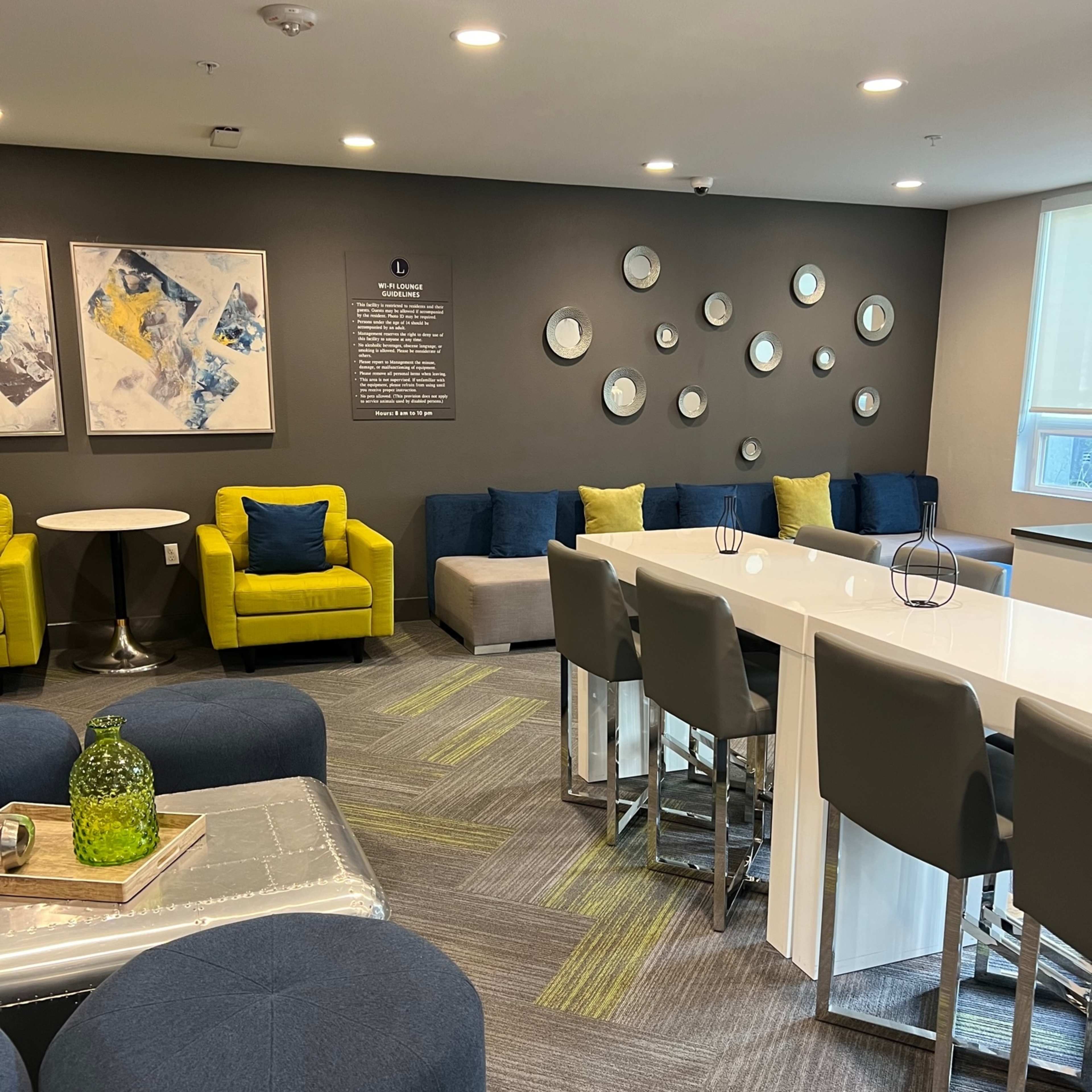 A modern lounge area with yellow and blue seating, a white communal table, and decorative wall mirrors.