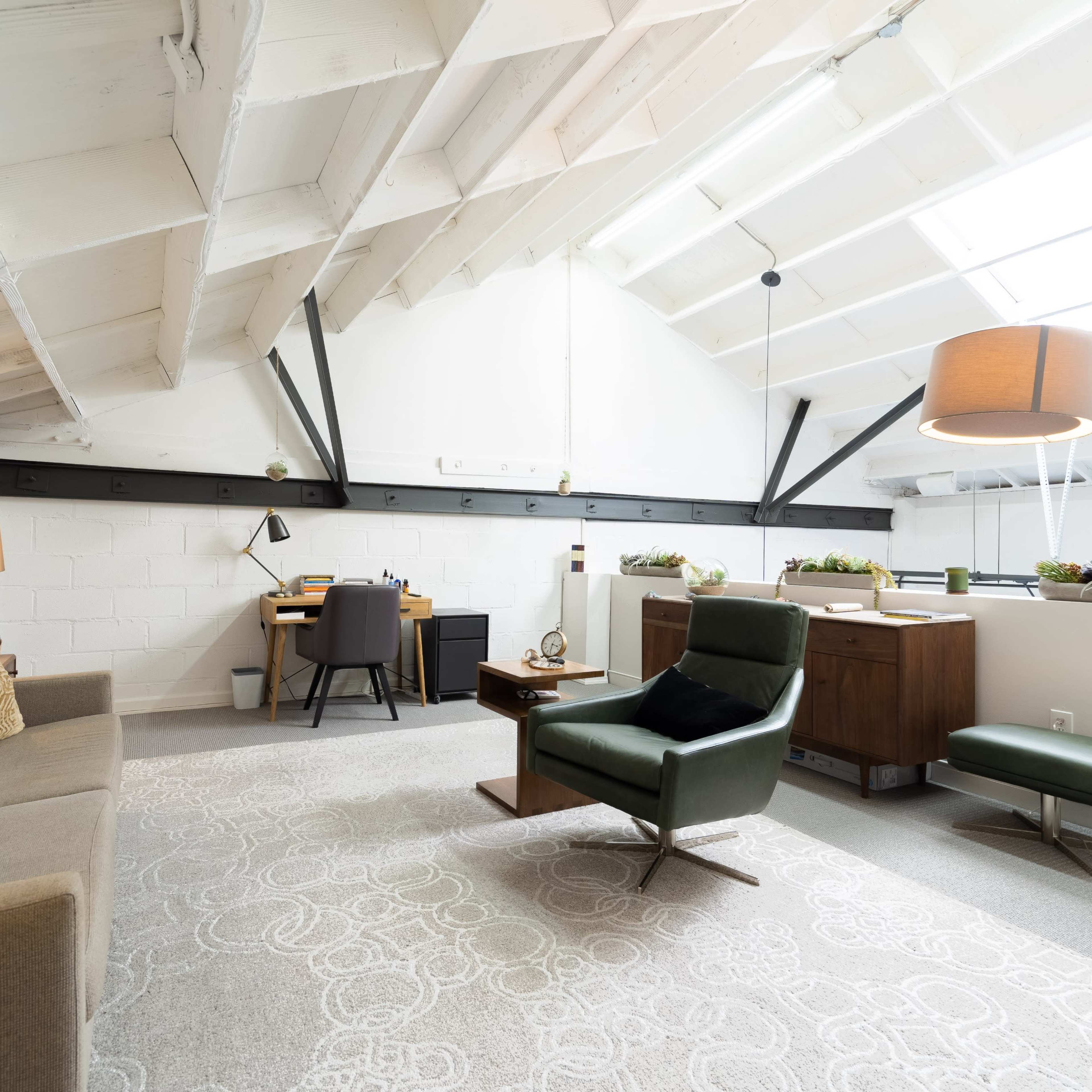A bright, modern workspace featuring a sofa, desk, and plants in a loft-style room with exposed beams and a skylight.