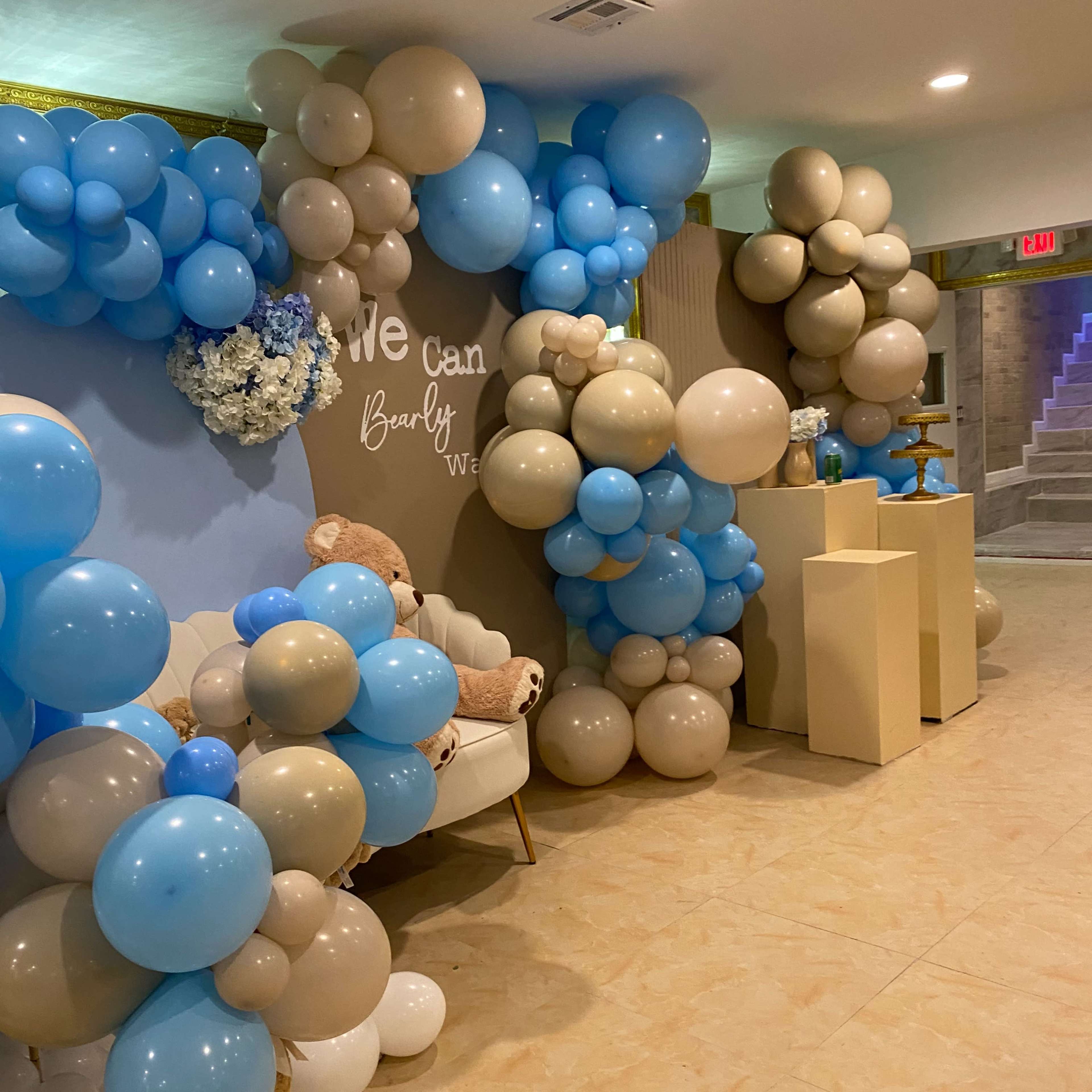 A decorated event space featuring a wall adorned with clusters of blue, beige, and white balloons, alongside a small seating area and decorative elements.