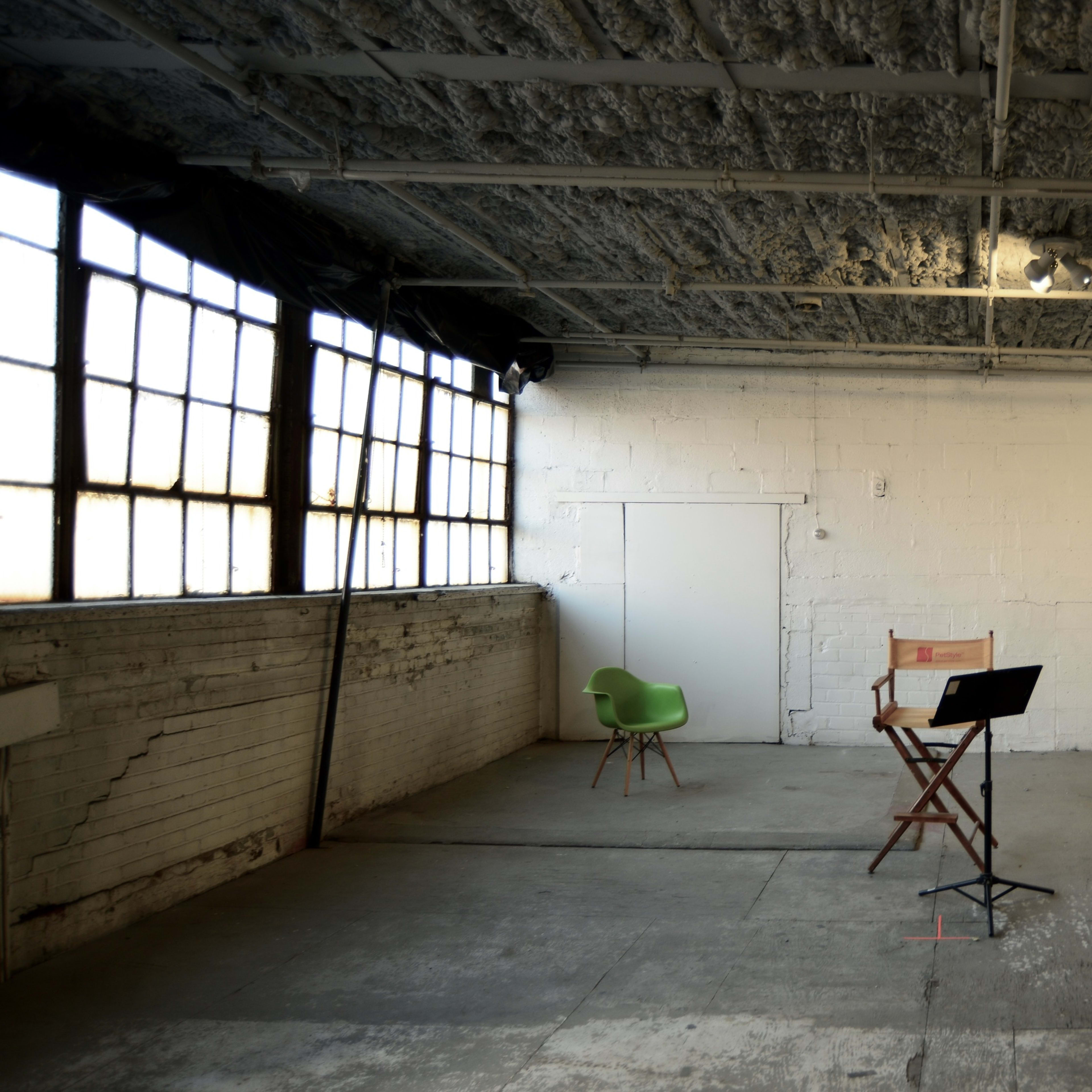 An empty industrial room with a wooden chair, a green chair, and a music stand on a concrete floor.