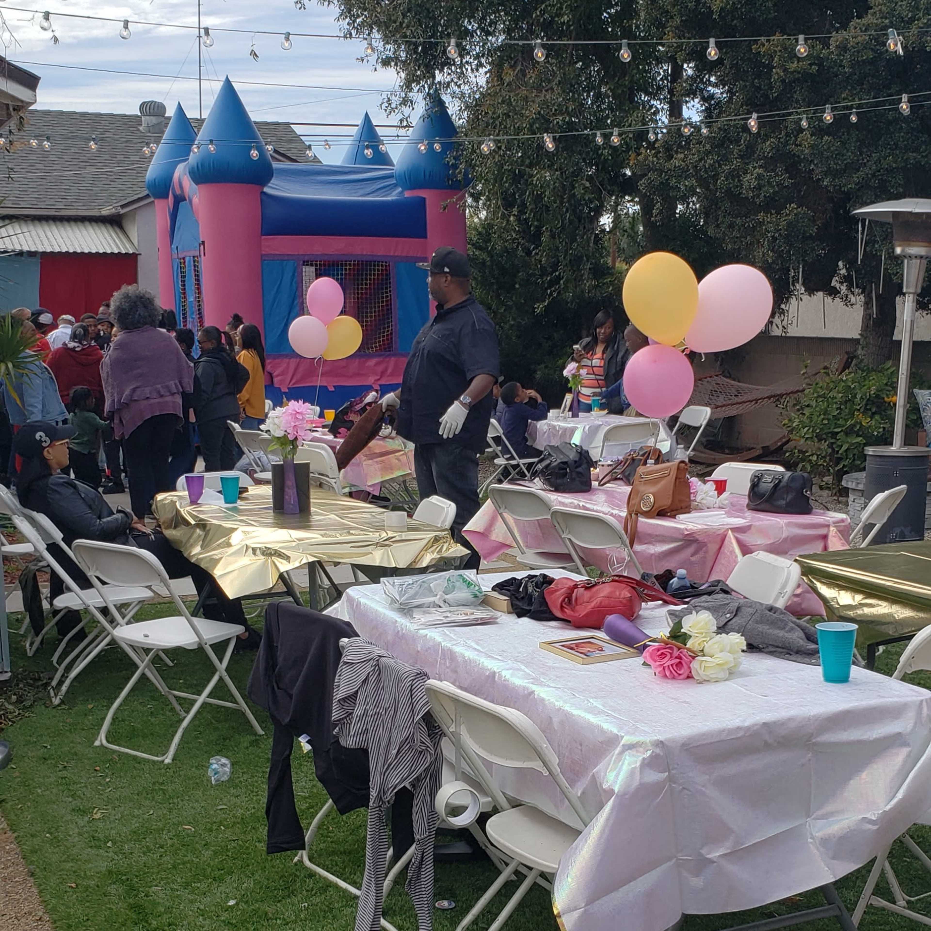 A backyard party is set up with tables covered in white tablecloths, balloons, and a bounce house in the background.