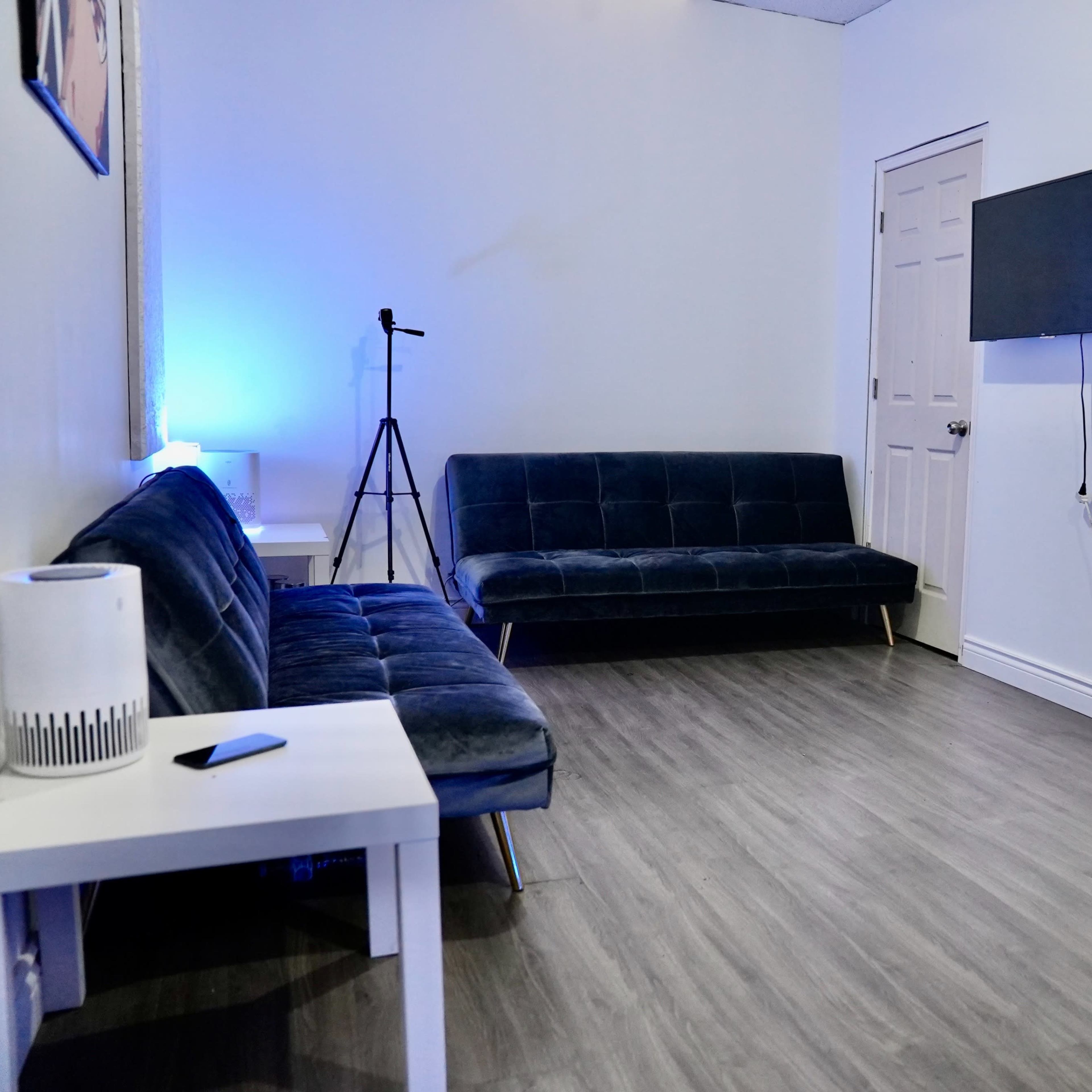 A minimalist room features two blue sofas, a small white table, a microphone stand, a wall-mounted TV, and a black office chair next to a keyboard.