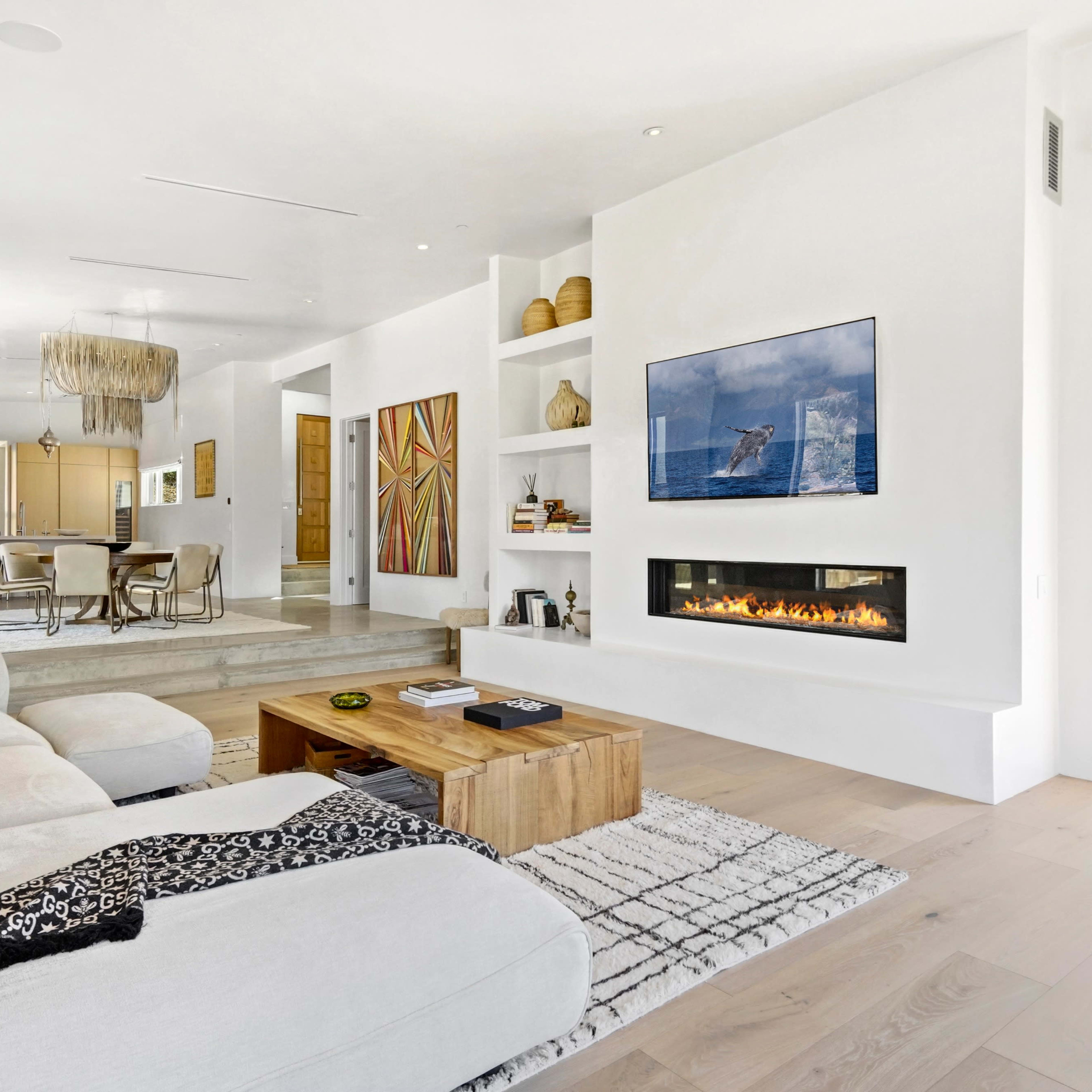 A modern living room features a white sectional sofa, a wooden coffee table, and a wall-mounted television above a linear fireplace, with large windows opening to an outdoor space.