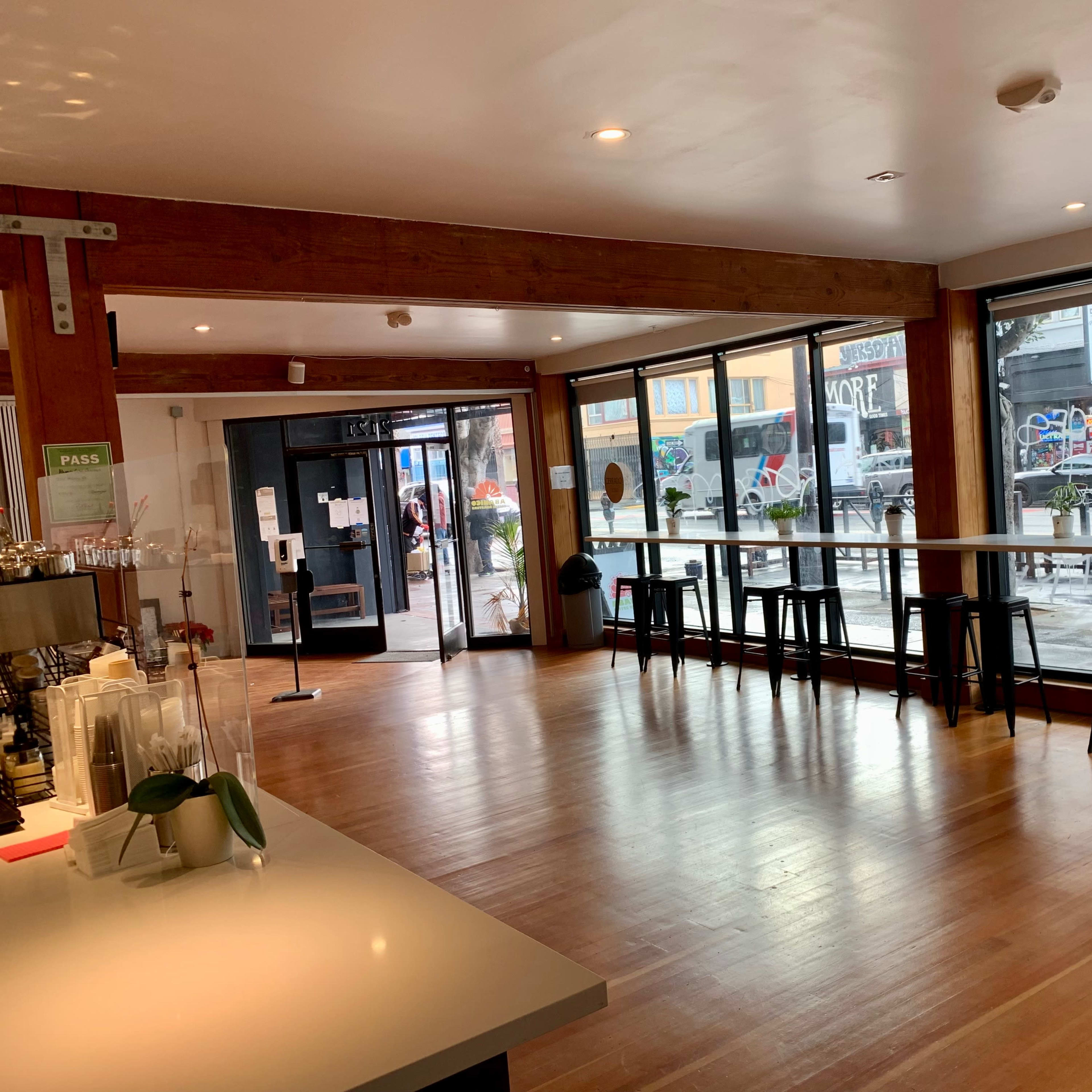An interior of a cafe with wooden beams, large windows allowing a view of a street with graffiti, and black high stools arranged near a long, white table.