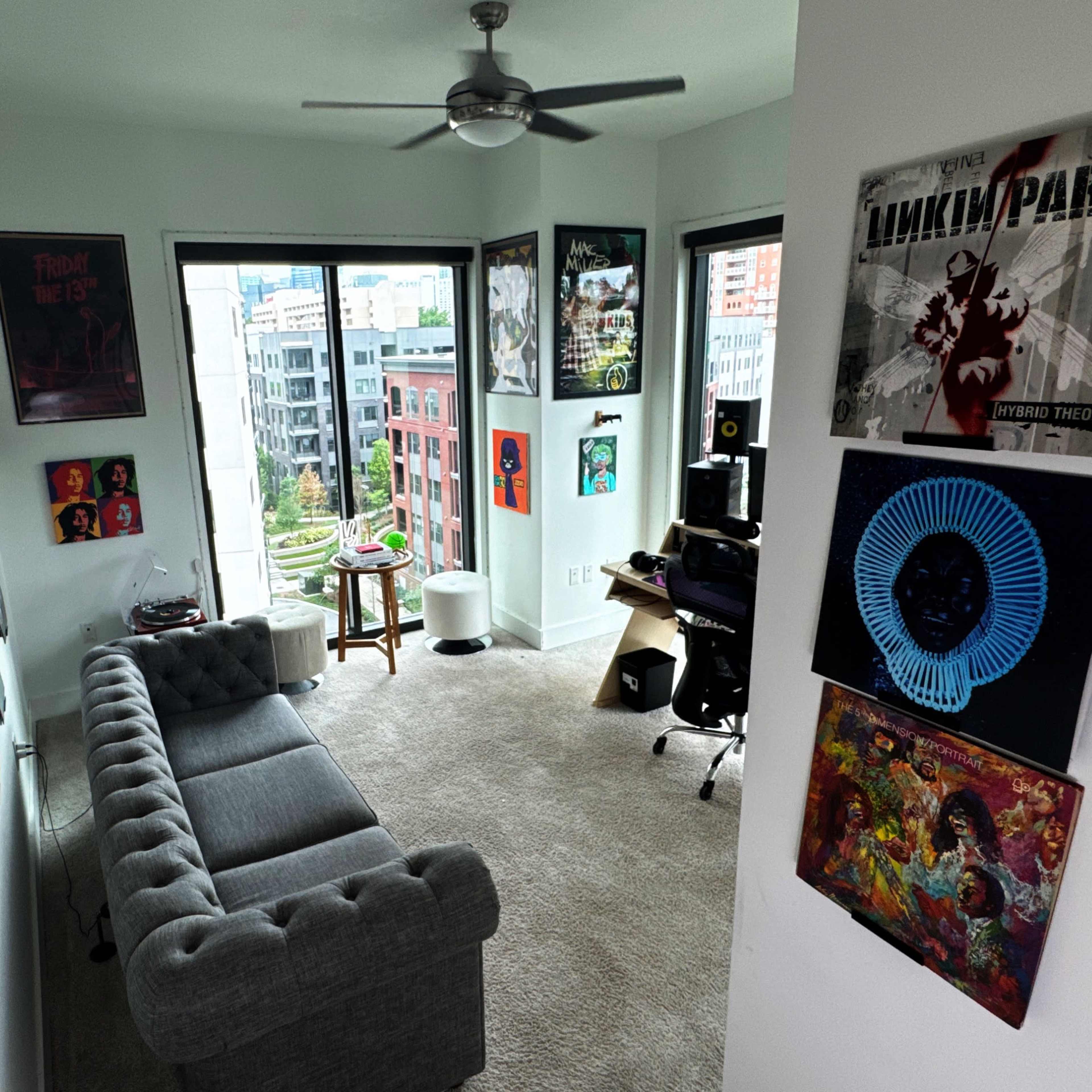 A cozy room with a gray couch, large windows overlooking a cityscape, and walls adorned with various posters and artwork.