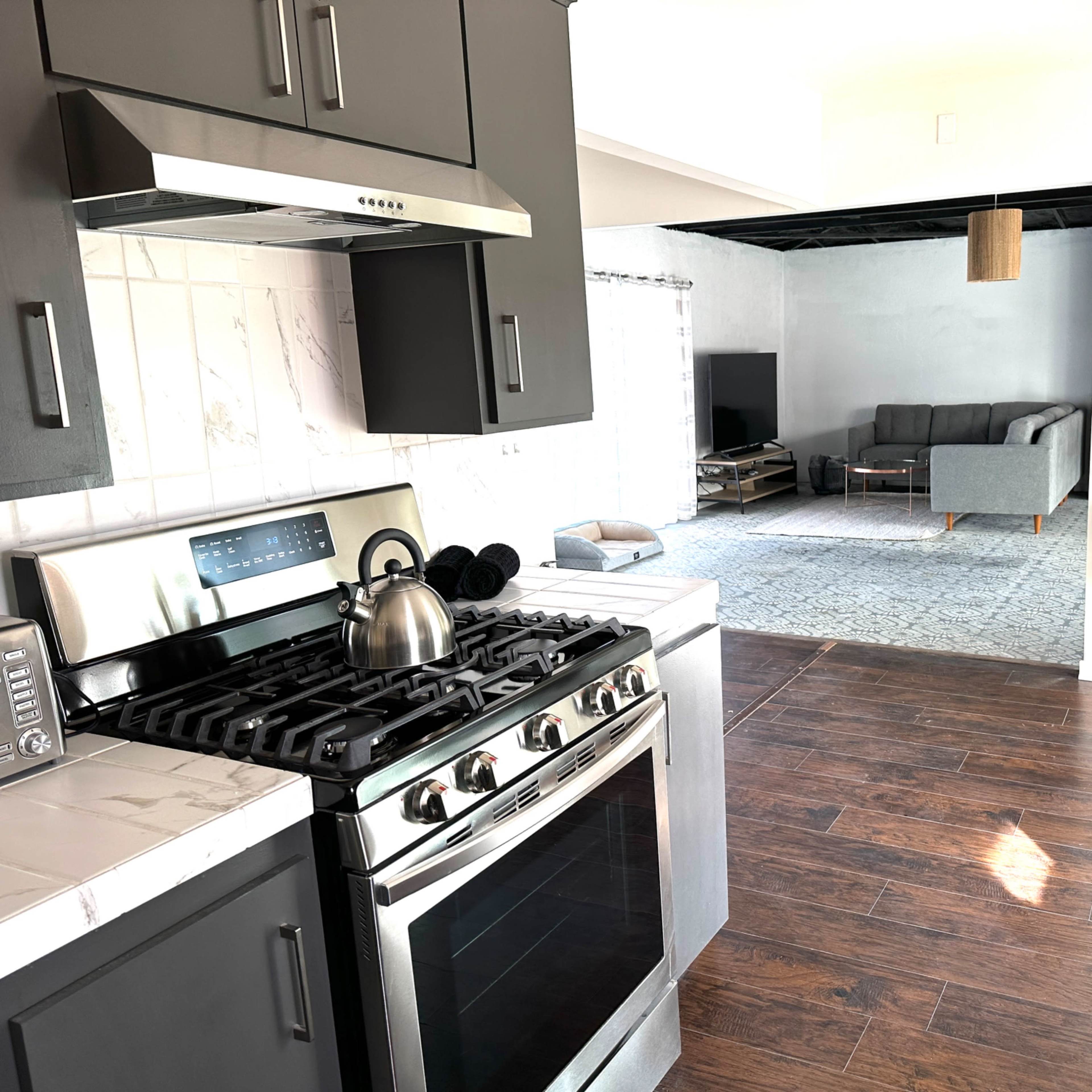 A kitchen with dark cabinetry, stainless steel appliances, and a view of a living area featuring a gray sofa and a small dining set in the background.