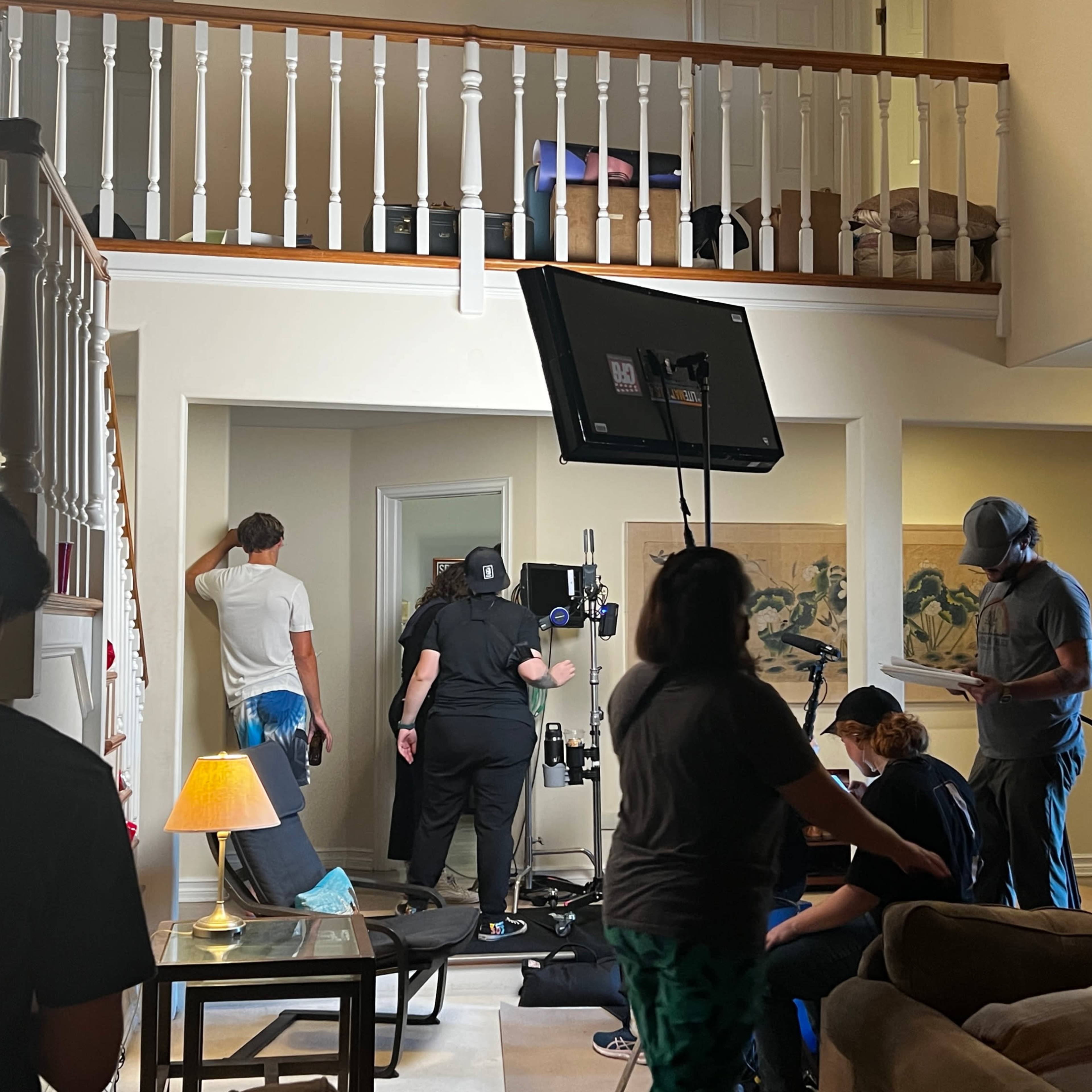 A film crew sets up equipment in a two-story living room, with various people preparing for a scene.