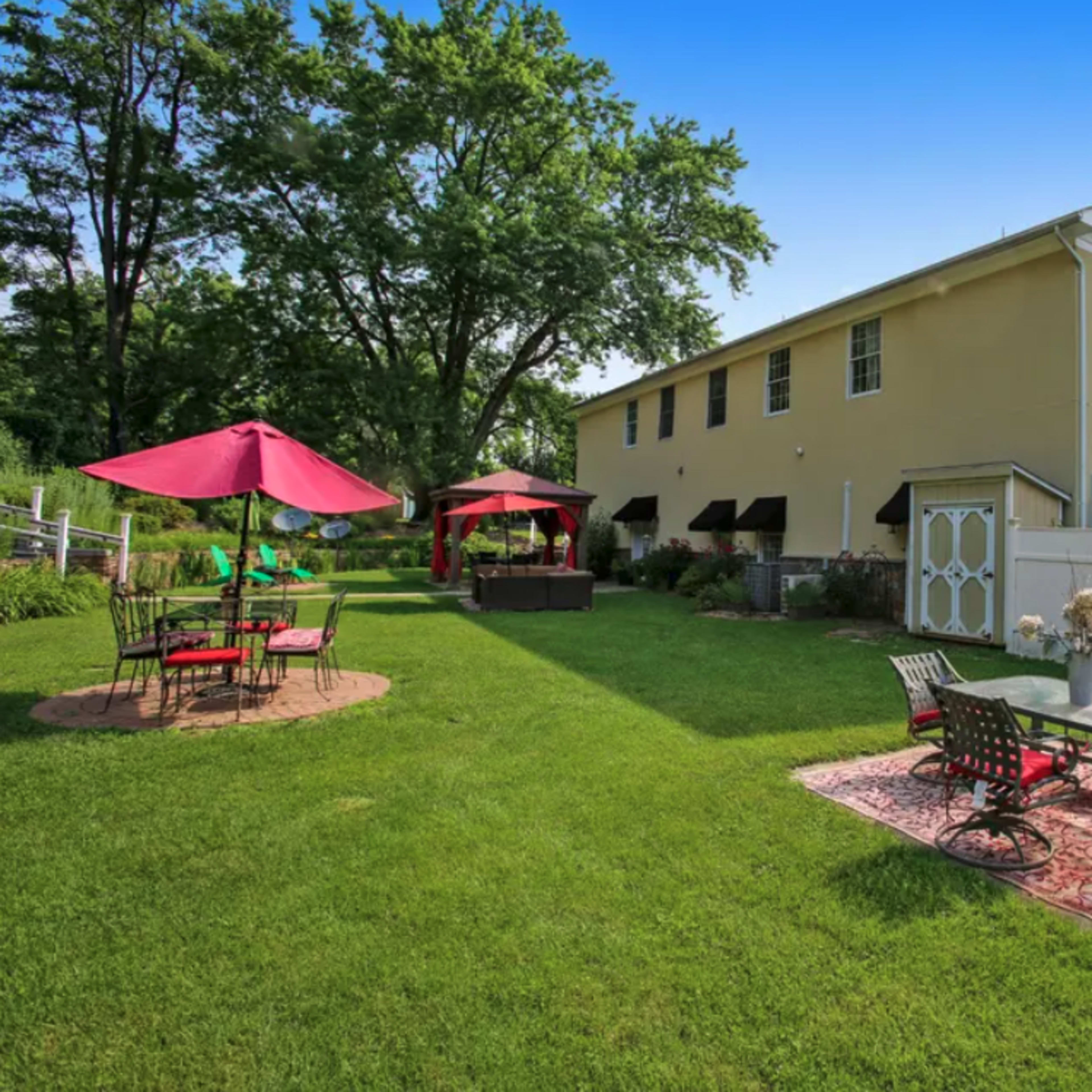 A spacious backyard featuring a beige house, a patio with a table and chairs, and two umbrellas over seating areas surrounded by green grass and trees.